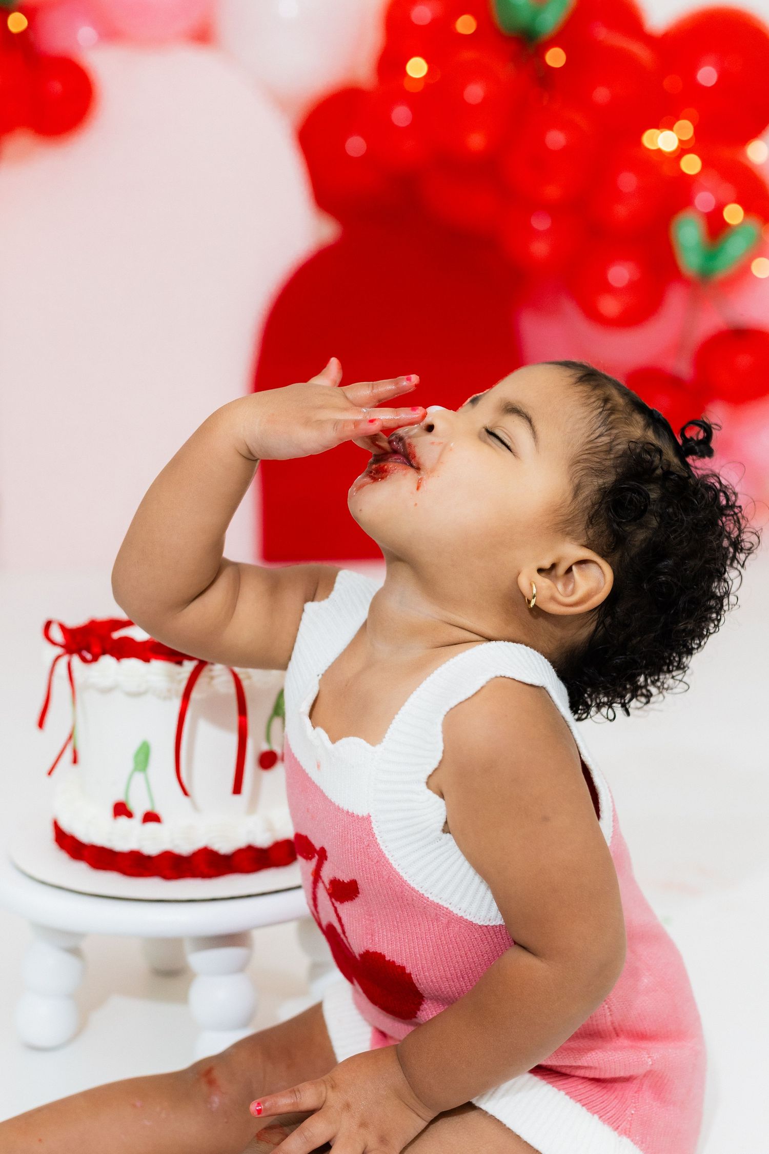 Toddler girl in pink dress enjoys smashing her cherry-themed first birthday cake at a red balloon party.
