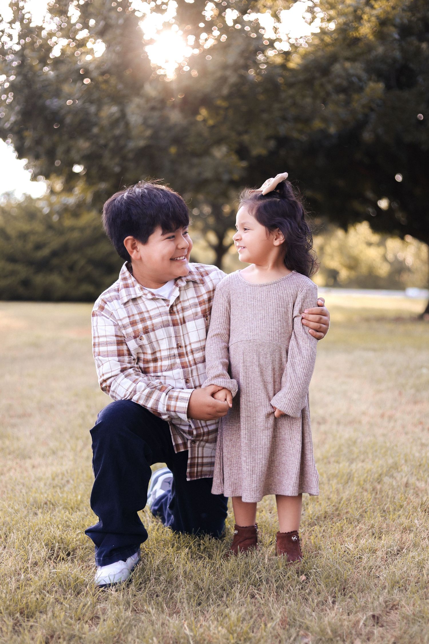 A boy kneels beside a little girl in a park, both smiling at each other in warm golden hour light.