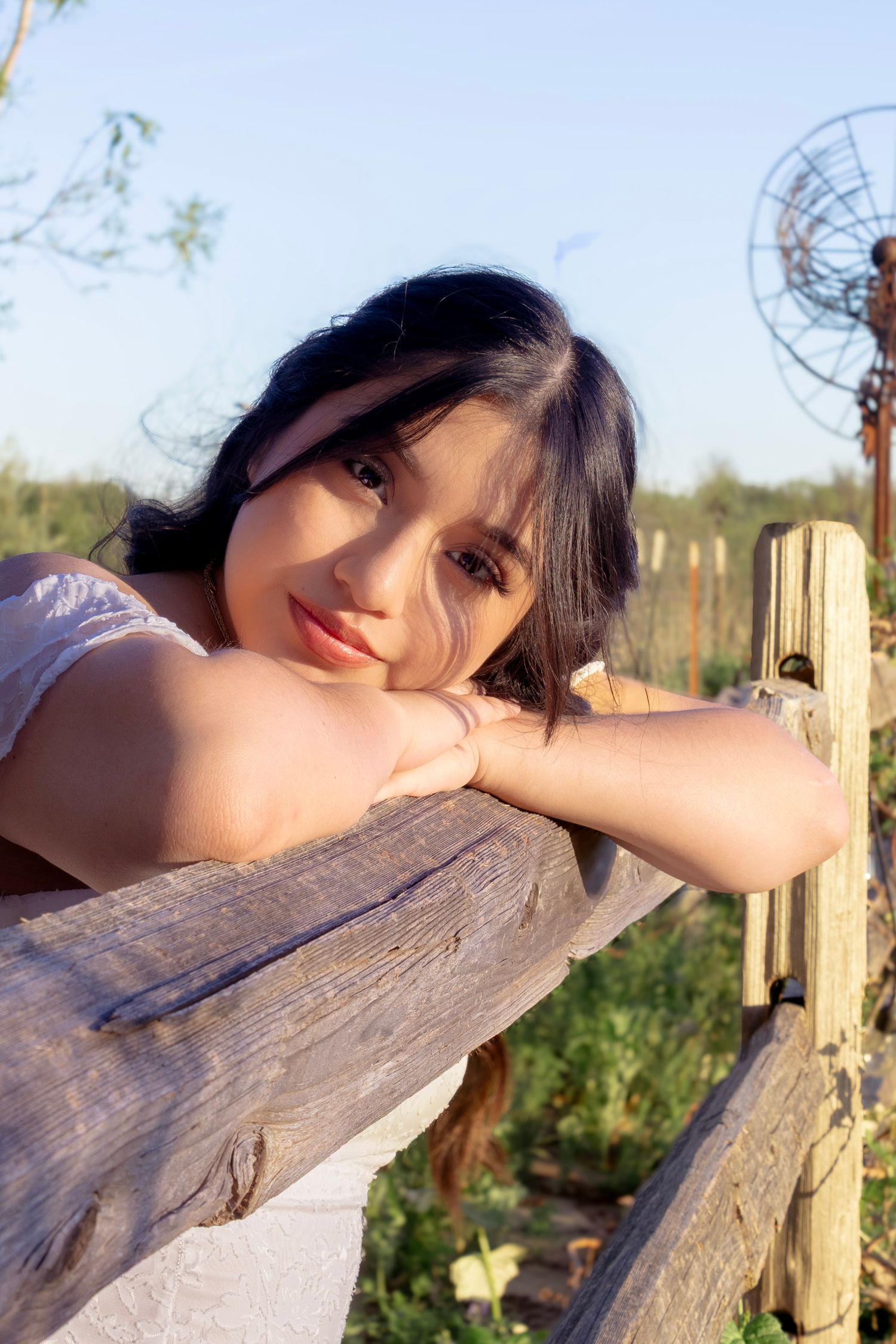 Young Asian woman smiling and leaning on a rustic wooden fence outdoors in warm sunlight.