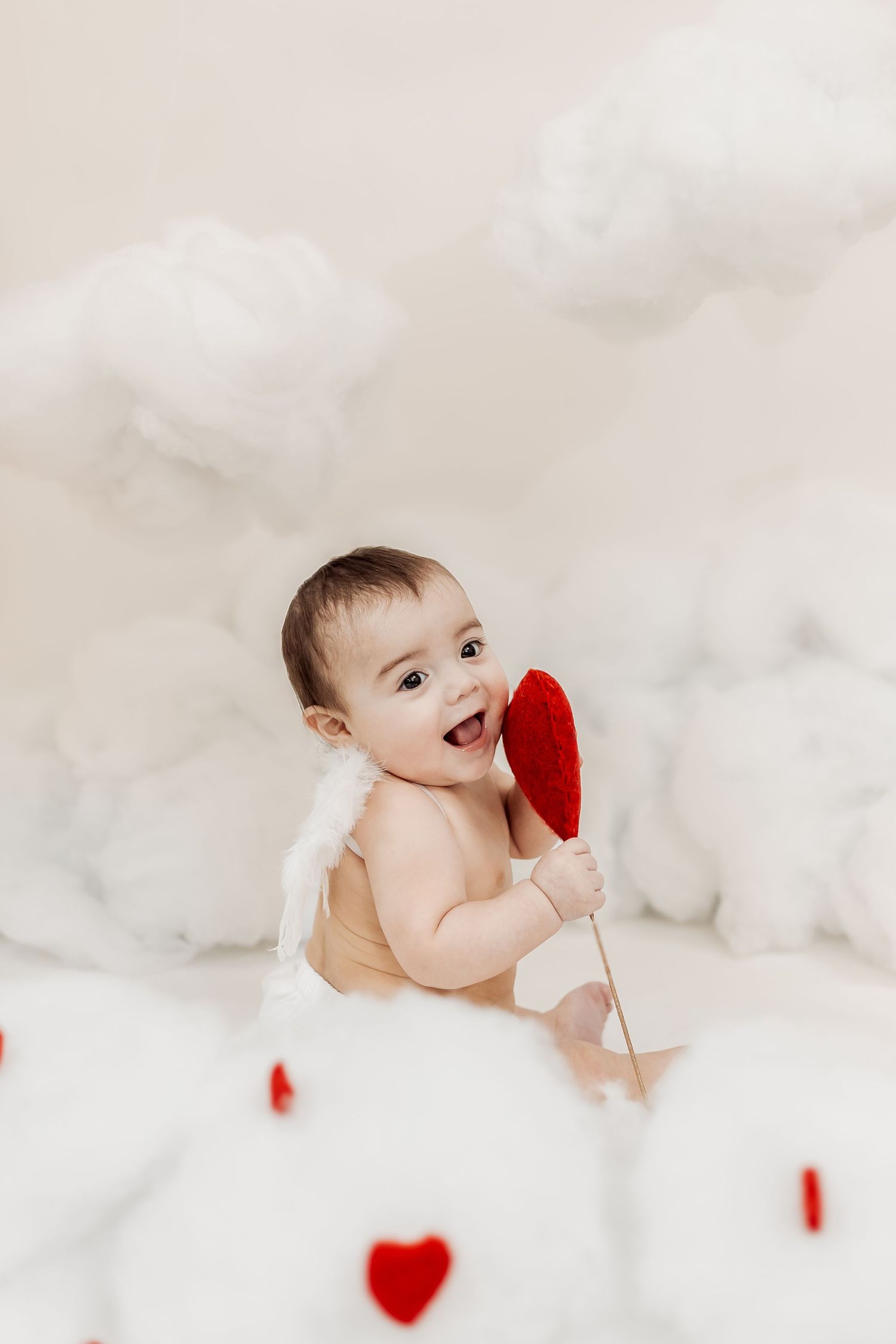 Joyful baby with angel wings holding a red heart arrow prop surrounded by white cloud decorations.