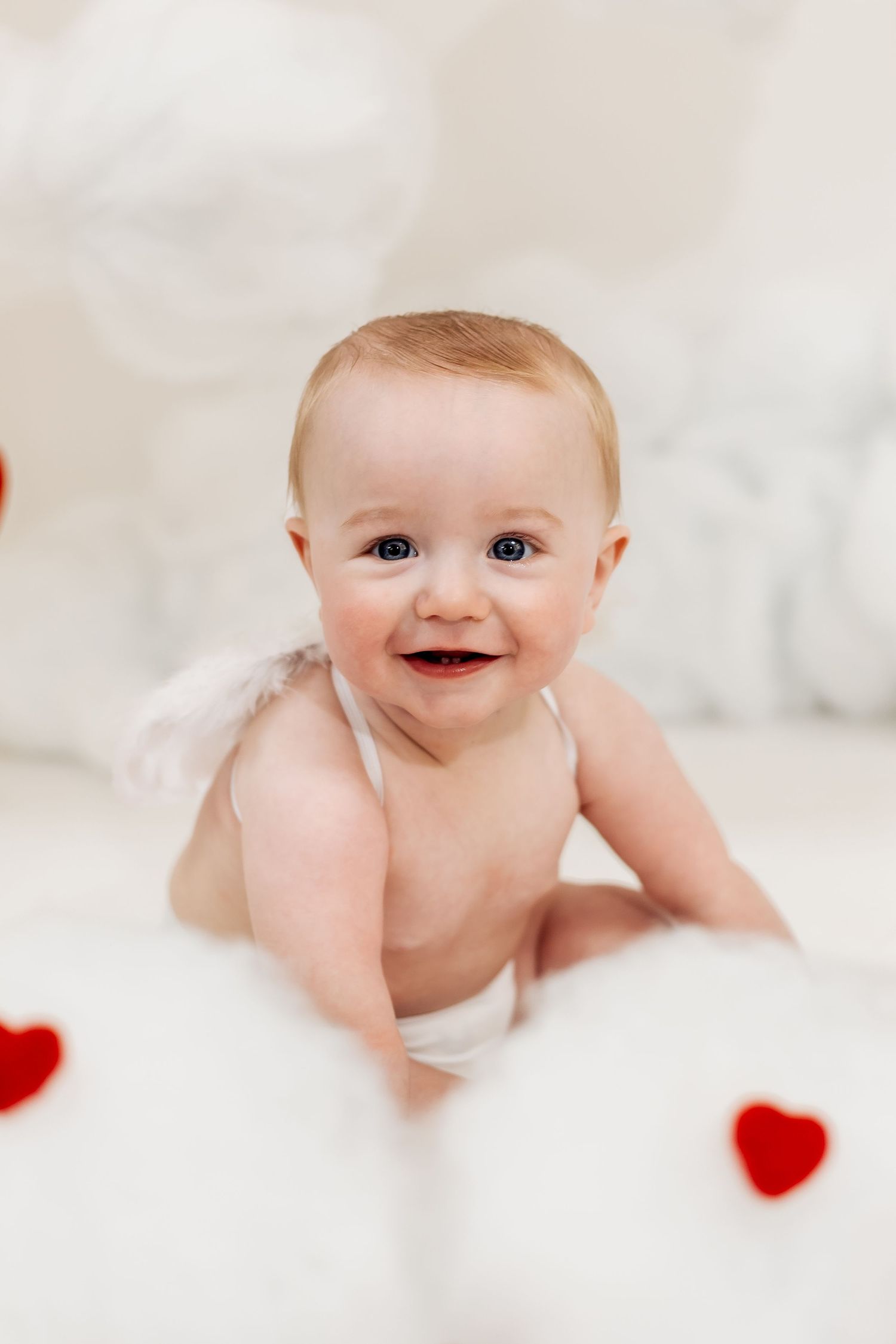 Adorable smiling baby dressed as Cupid with white wings surrounded by red hearts on a soft white background.