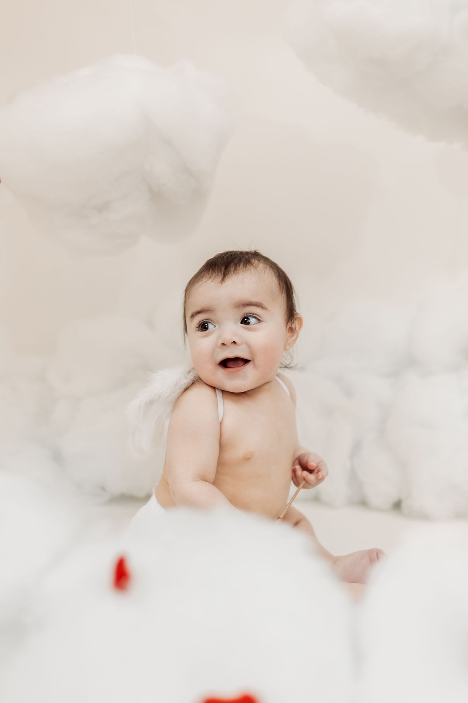 Cute baby with angel wings sitting among fluffy white cloud decorations, looking up with a smile.
