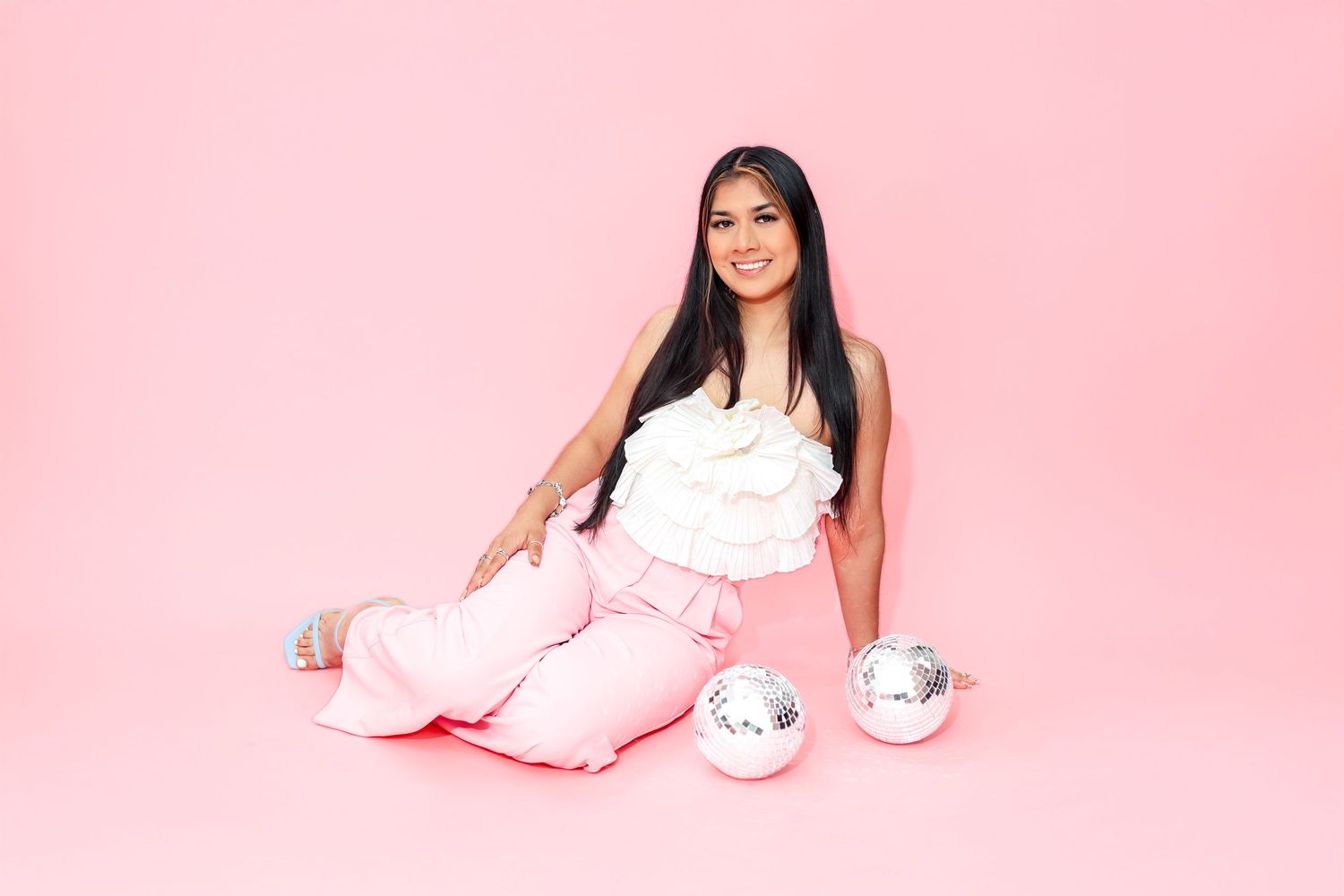 A young woman in pink pants and white top sits cross-legged on a pink backdrop with silver disco balls.