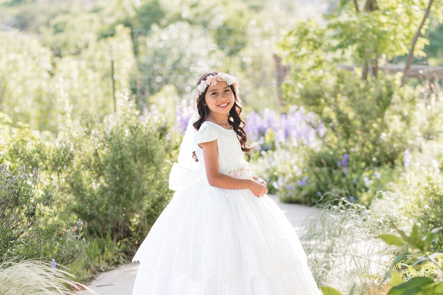 Young girl in white dress and floral crown smiles in a sunlit garden with purple flowers.