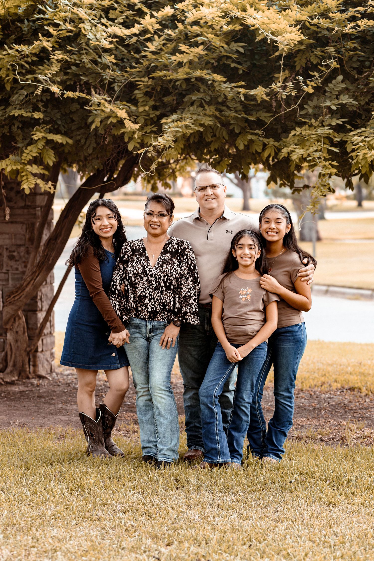 Family of five smiling together outdoors under a large tree on a sunny day.