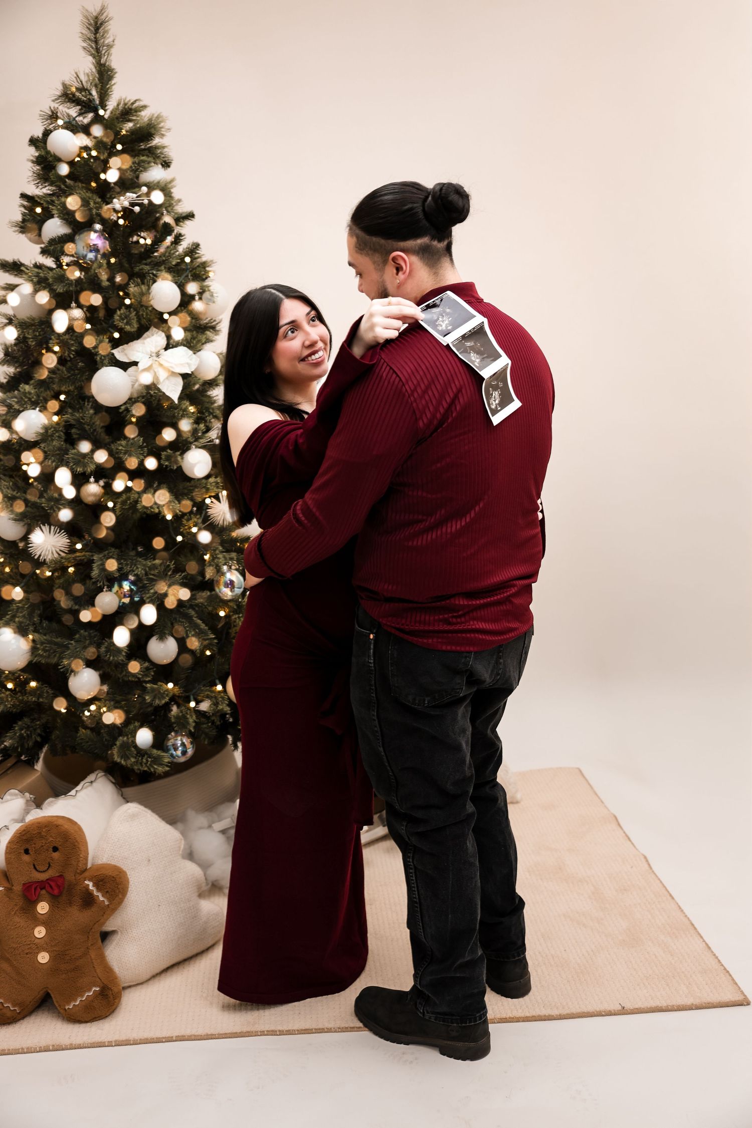 Couple in burgundy outfits embracing near decorated Christmas tree with ultrasound photos on his back.