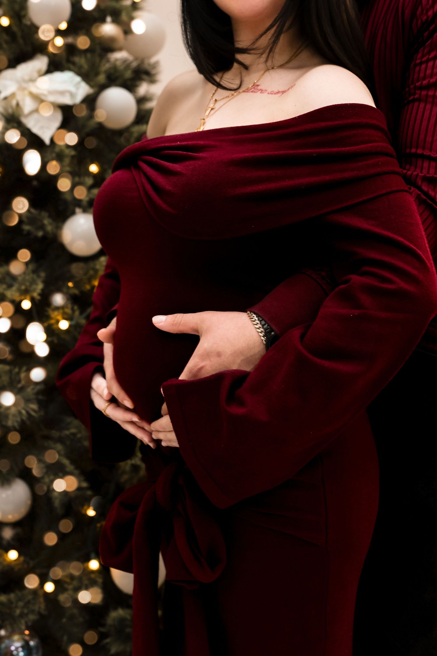 Close-up of pregnant belly with couple's hands cradling it in front of a glowing Christmas tree.