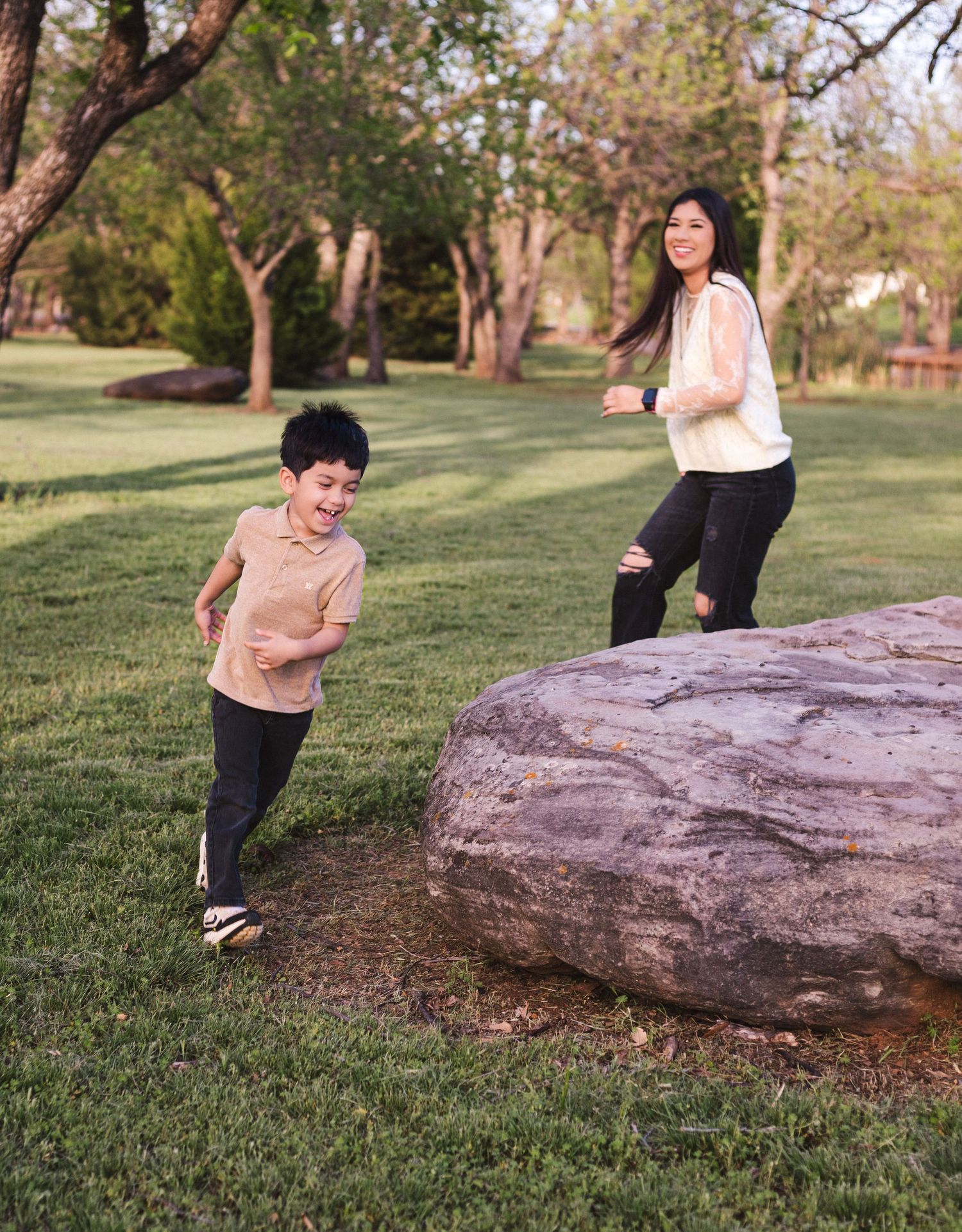 A joyful Asian mother and son playing and running around a large rock in a lush green park.