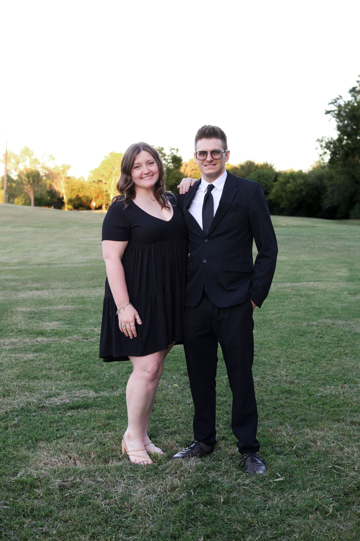 Smiling couple in matching black outfits posing on green grass at sunset outdoors.
