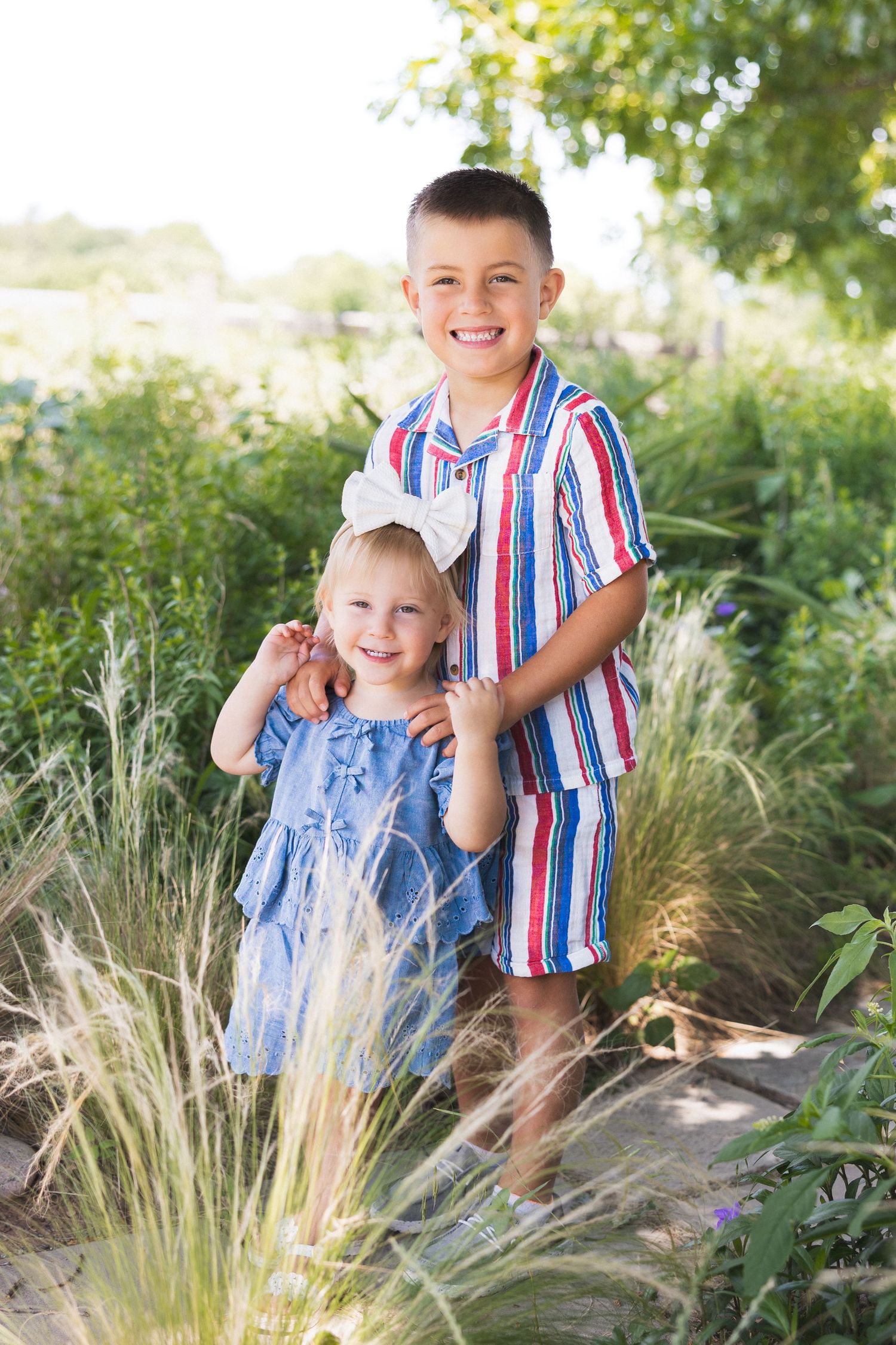 A smiling boy in a striped outfit stands with his little sister in a sunlit garden path.