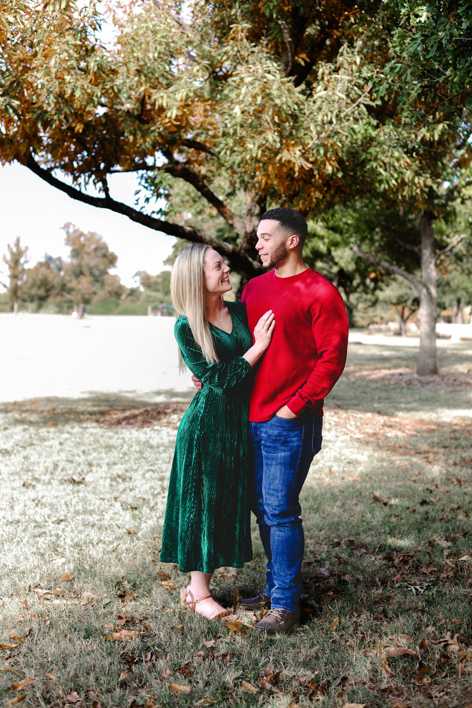 A couple in red and green outfits poses together under autumn trees in an outdoor setting.