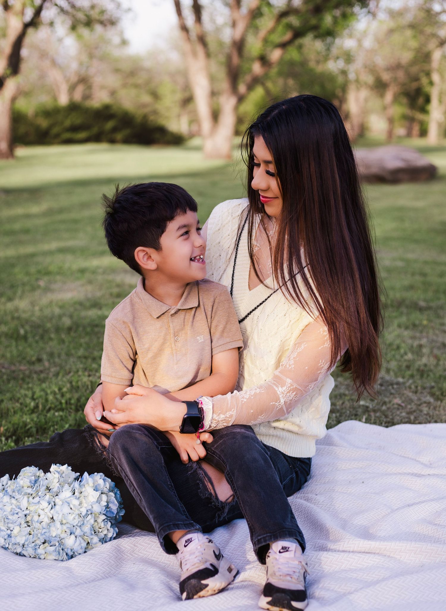 A woman and young boy share a joyful moment on a blanket in a park, surrounded by blue hydrangeas.