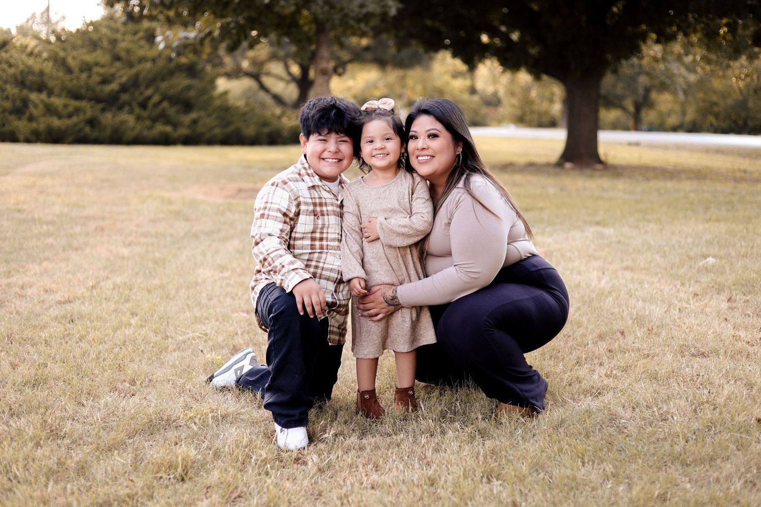 A mother crouches in a grassy park with her young son and toddler daughter for a family portrait.