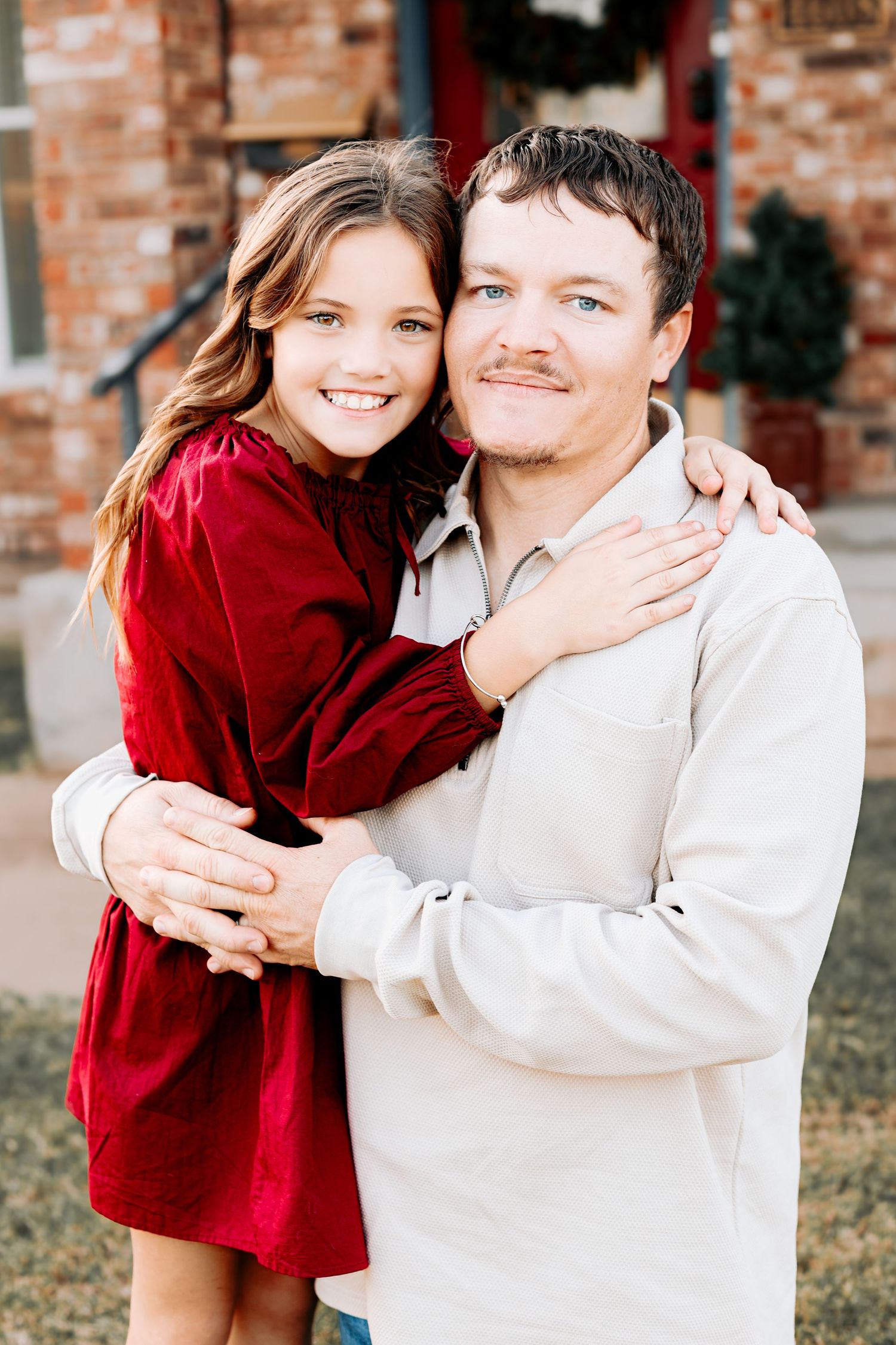 A young girl in a red dress hugs a man in a beige shirt while both smile at the camera outdoors.