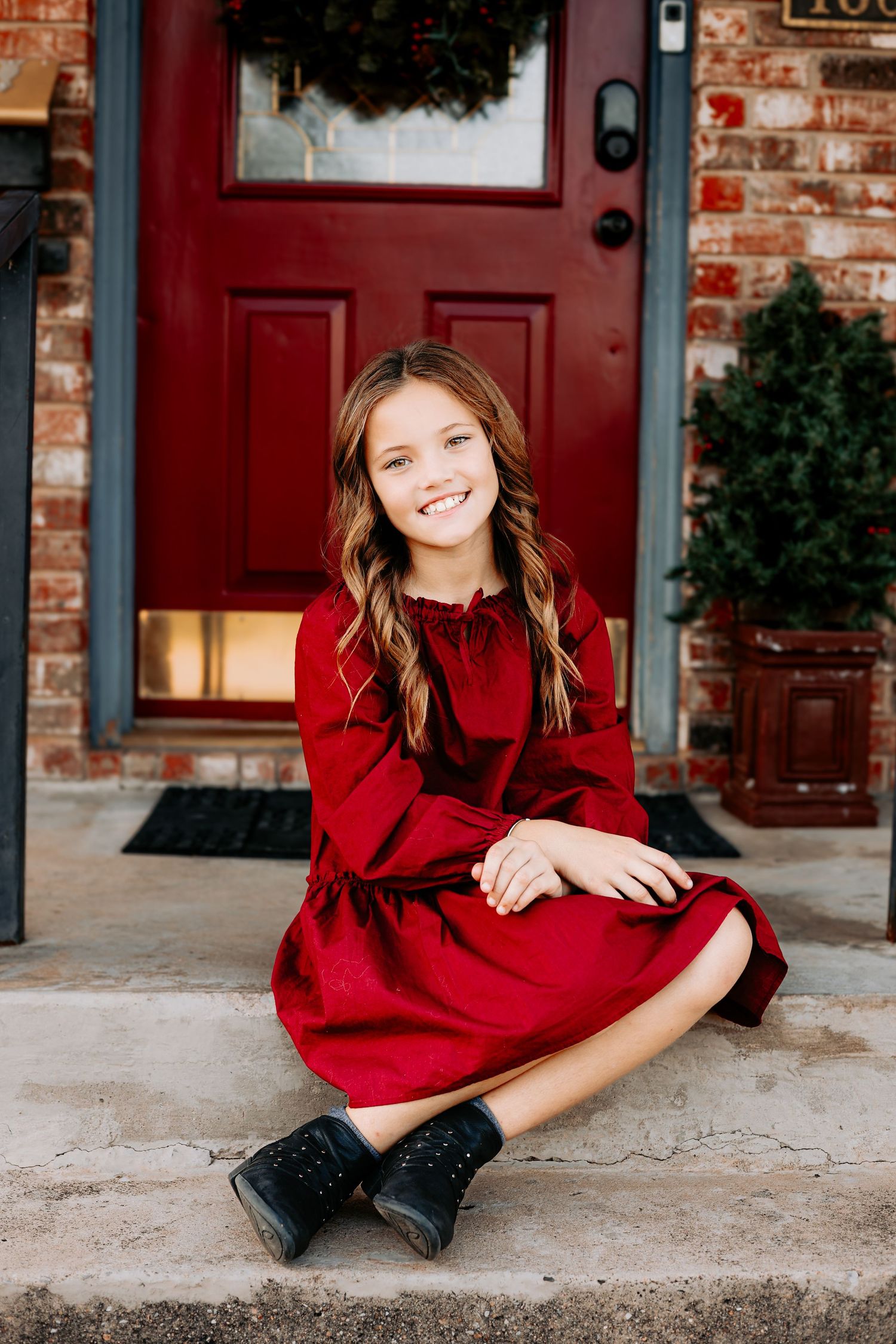 A young girl in a red dress and black boots sits on front porch steps smiling at the camera.