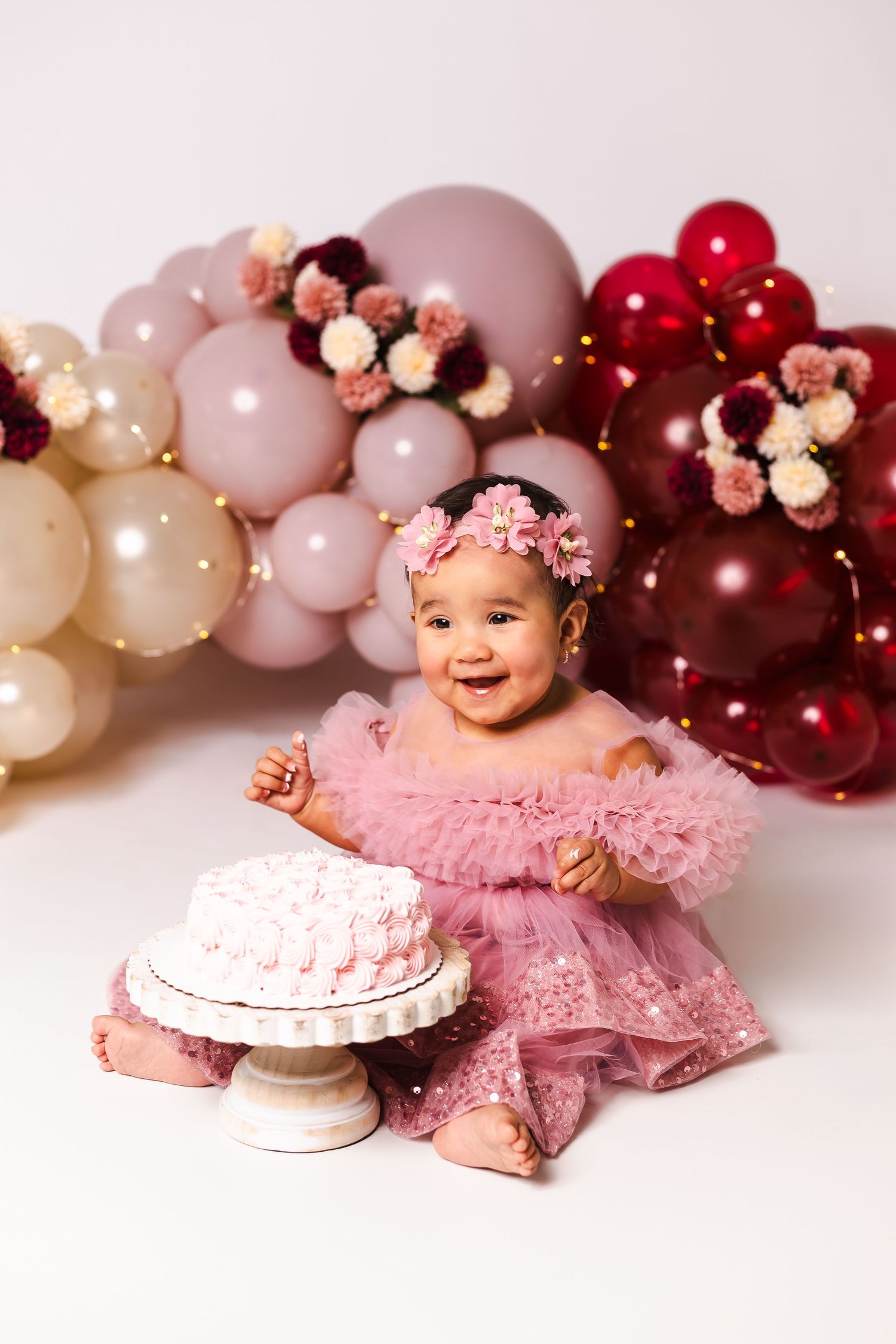 Adorable baby girl in pink tutu dress celebrates first birthday with cake and balloon backdrop.