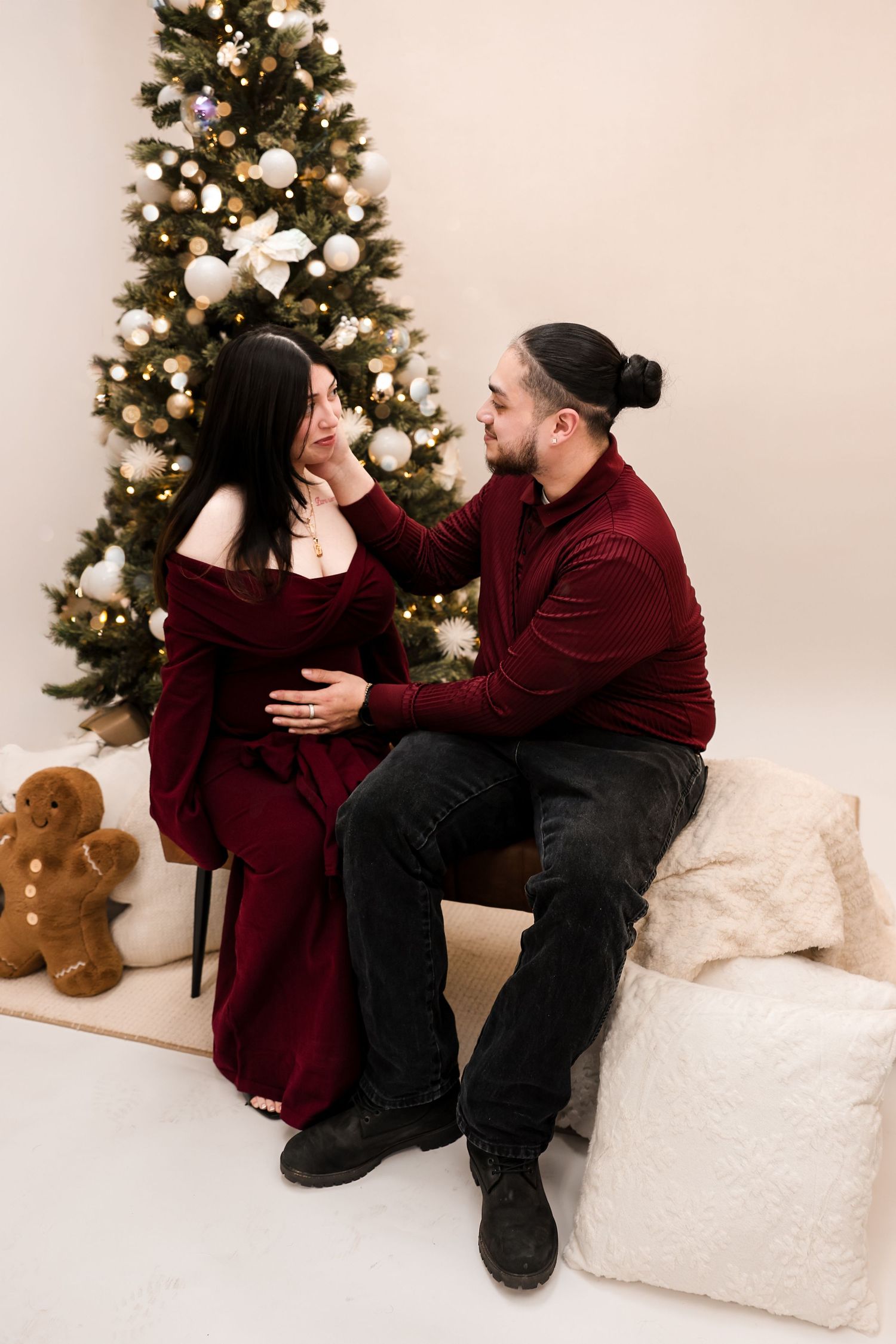 Couple in burgundy outfits sit together gazing lovingly at each other beside a decorated Christmas tree.