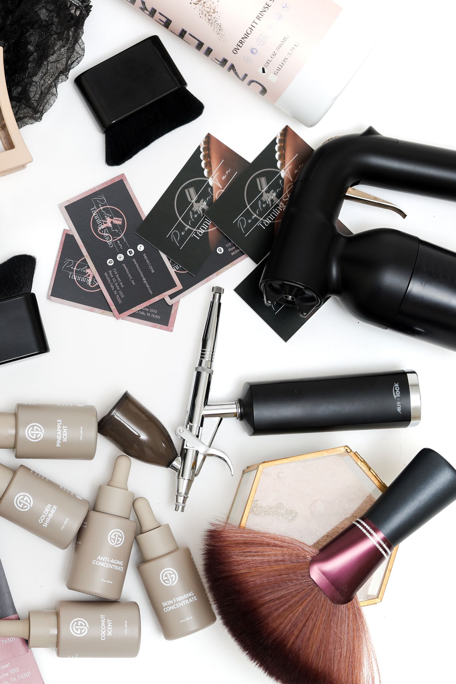 Flat lay of airbrush makeup tools, business cards, foundation bottles, and a kabuki brush on white background.