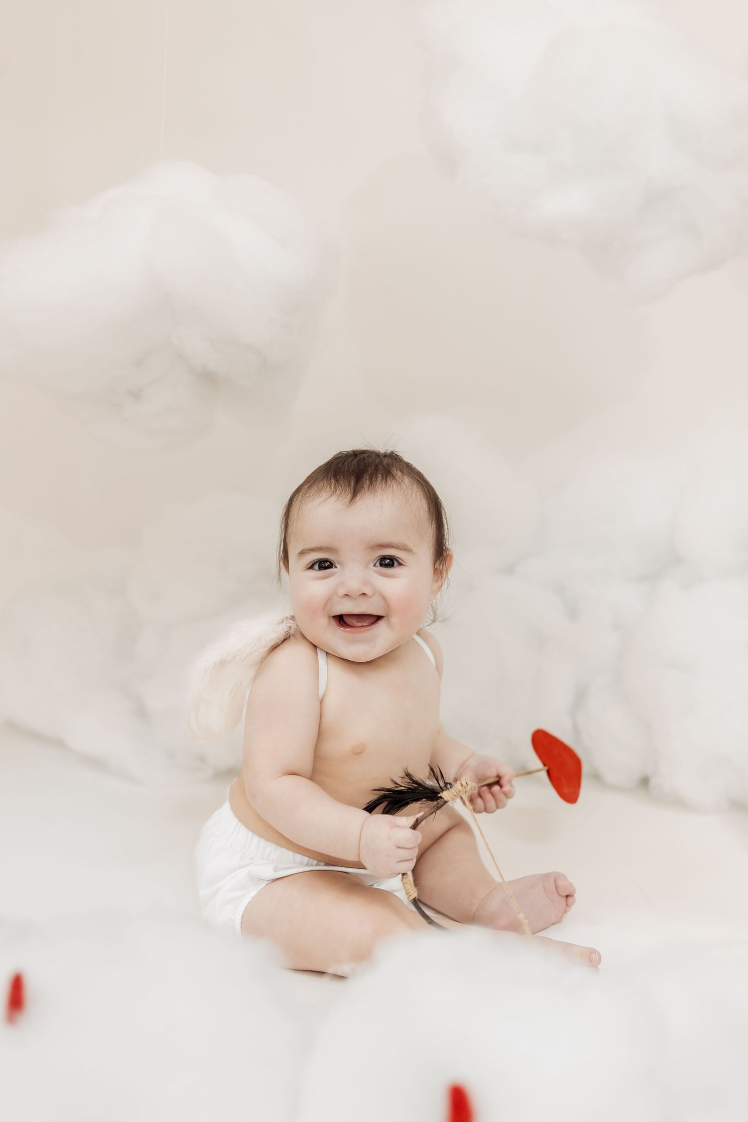 Smiling baby in diaper with white angel wings holding a red heart arrow among white cloud props.