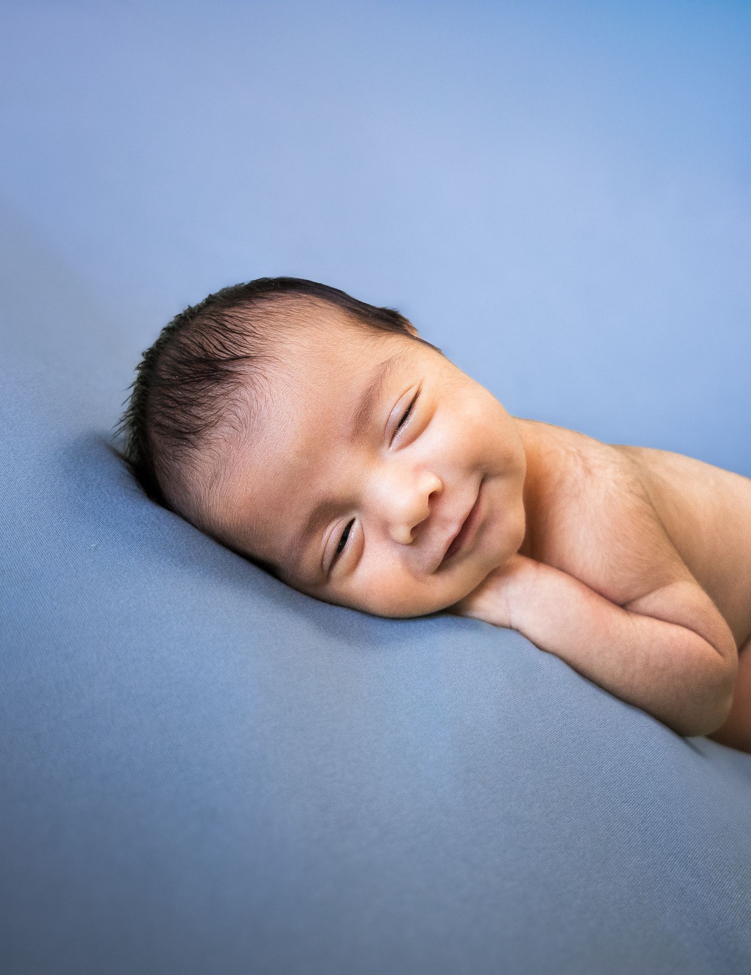 Close-up of smiling newborn baby with dark hair resting on blue backdrop.