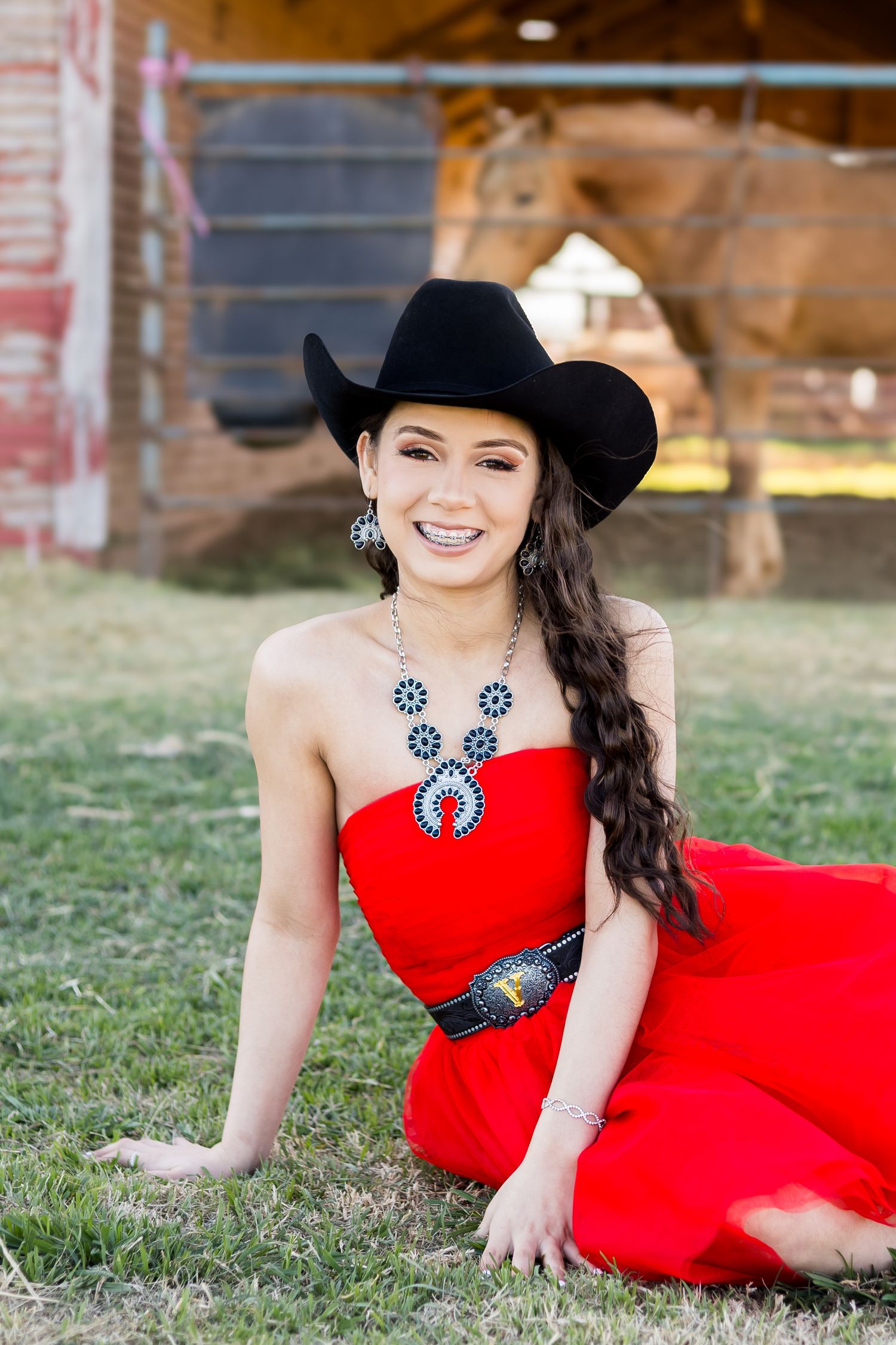 Young woman in red strapless dress, black cowboy hat, and turquoise jewelry sits on grass near a barn.