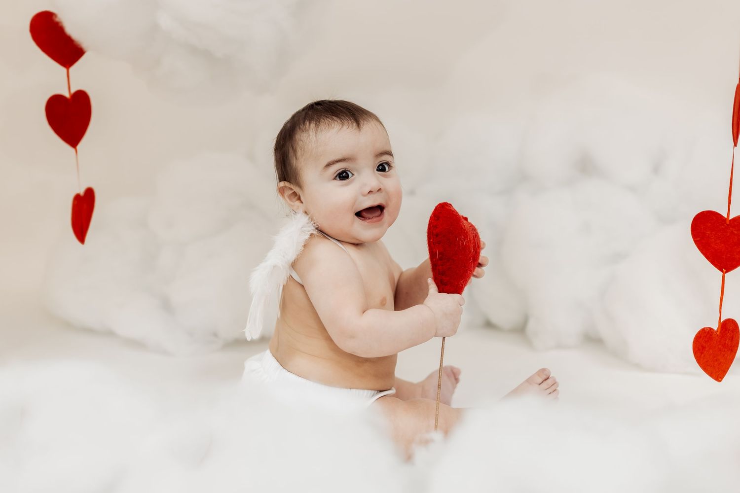 Adorable baby in diaper with angel wings holds red heart prop surrounded by Valentine's Day heart decorations.