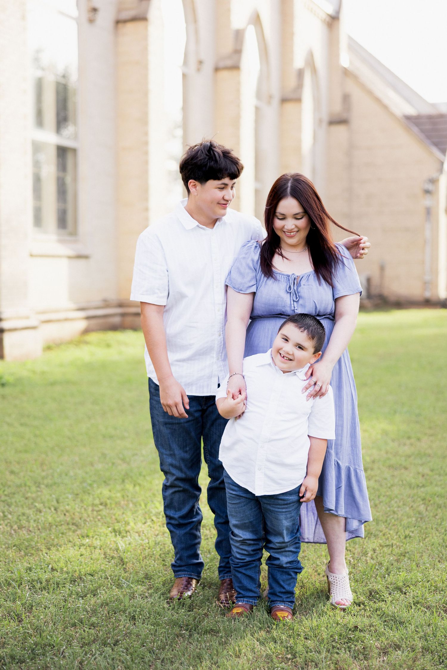 Happy family of three with pregnant mother in blue dress poses outdoors near a historic building.