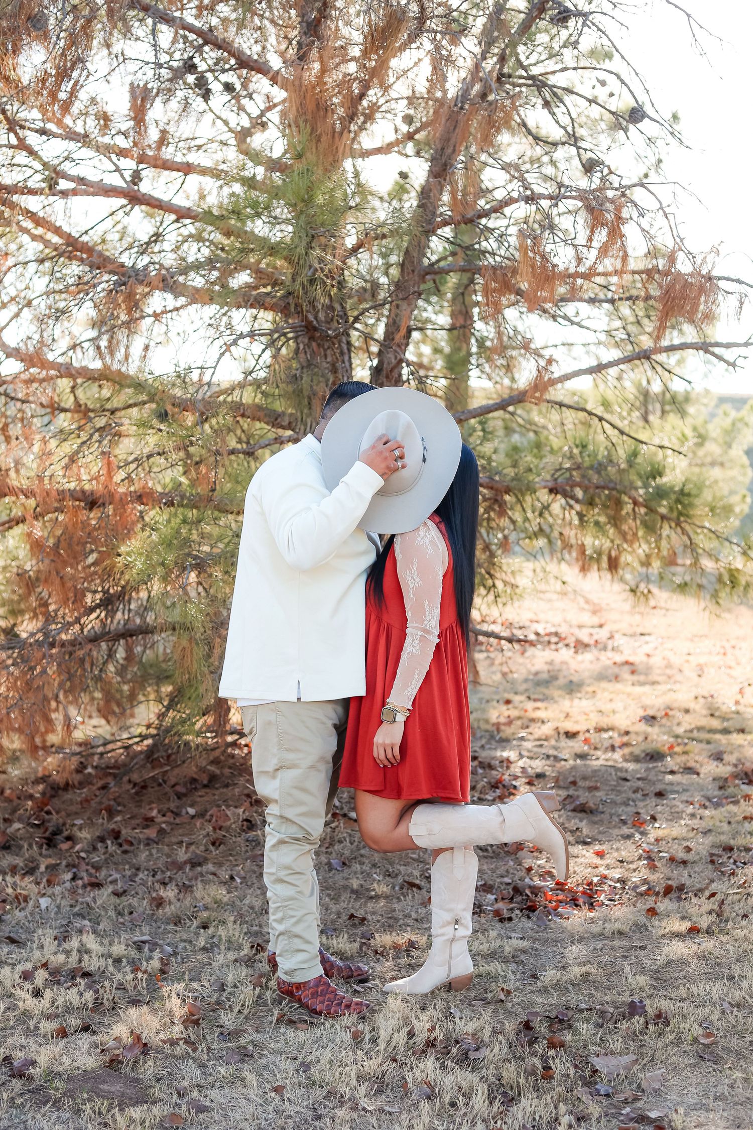 Couple sharing a kiss hidden behind a cowboy hat outdoors near a pine tree in matching outfits.
