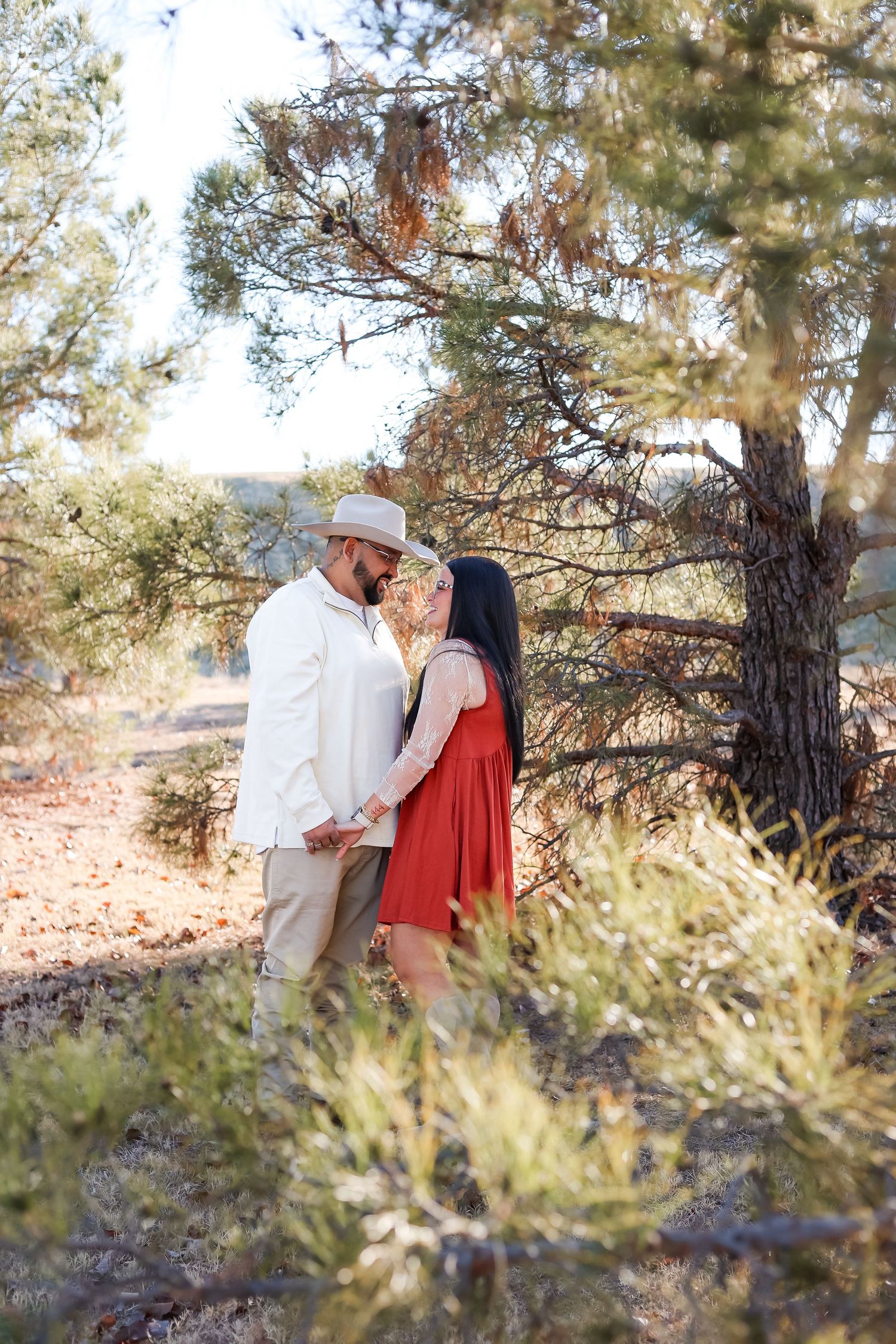 Couple sharing a romantic moment holding hands among pine trees, man in white shirt and hat, woman in red dress.