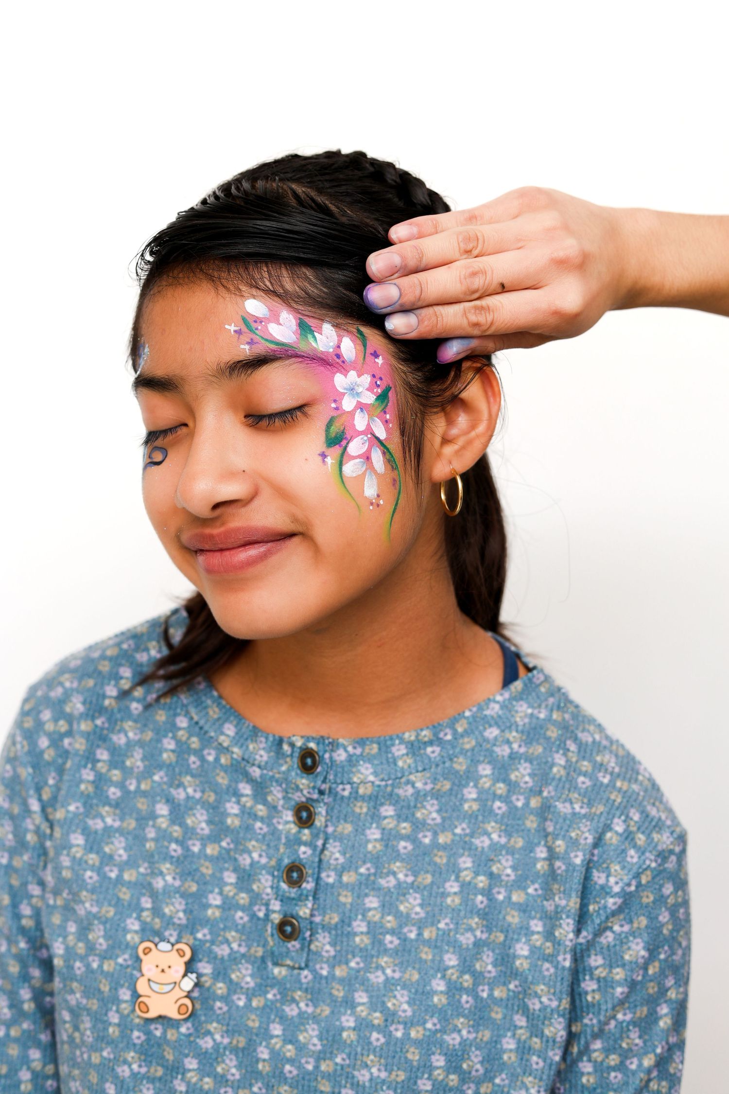 Young girl with floral face paint design being applied by a face painting artist on white background.