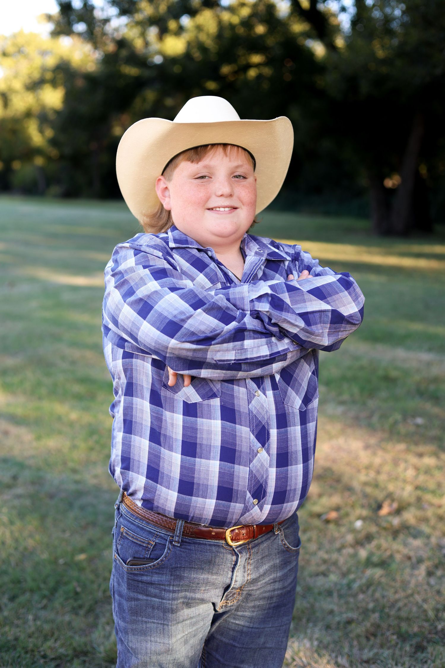 Smiling red-haired boy in cowboy hat and plaid shirt posing with arms crossed outdoors.