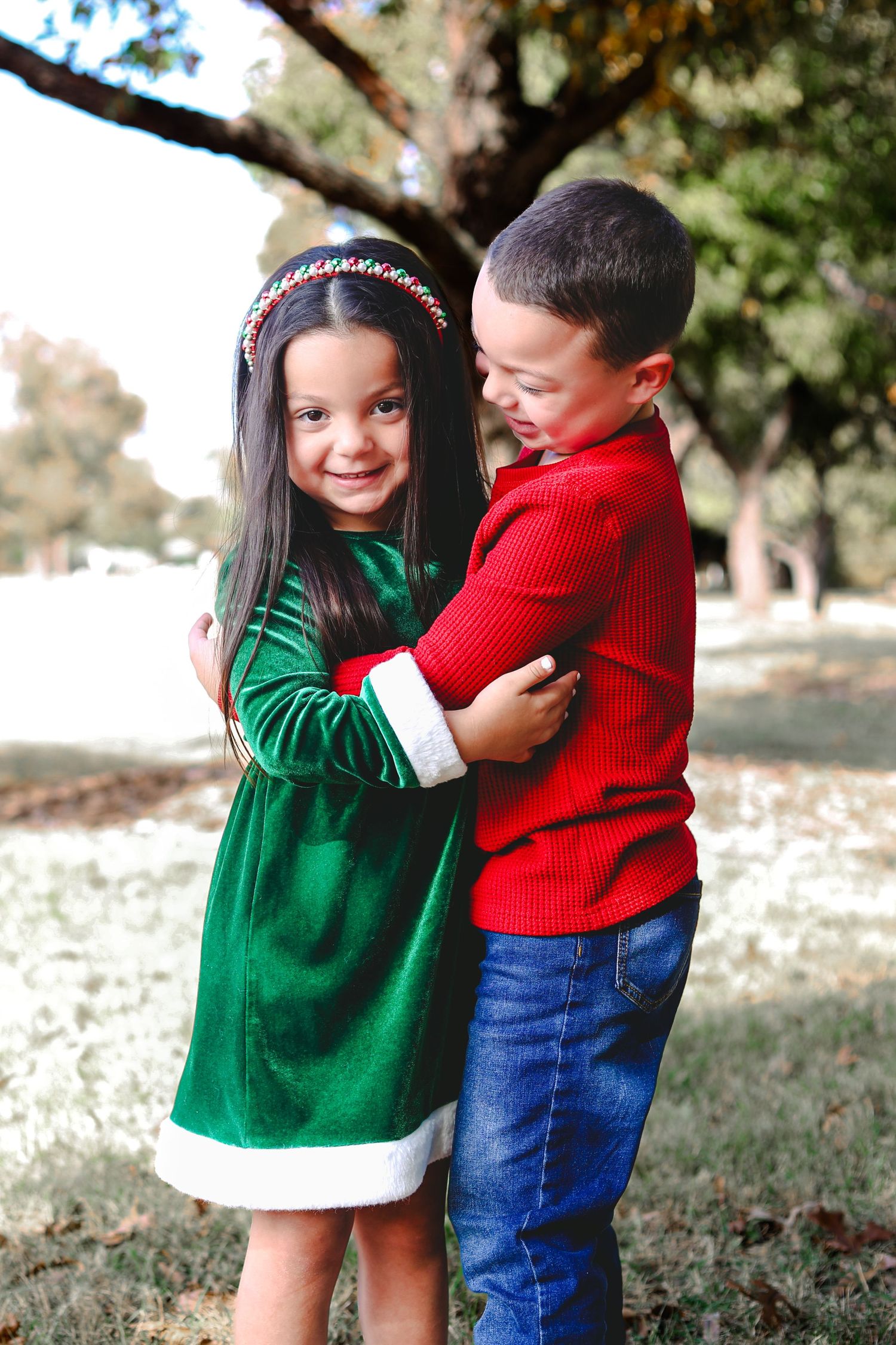 Two children in Christmas outfits hugging outdoors, girl in green velvet dress and boy in red sweater.