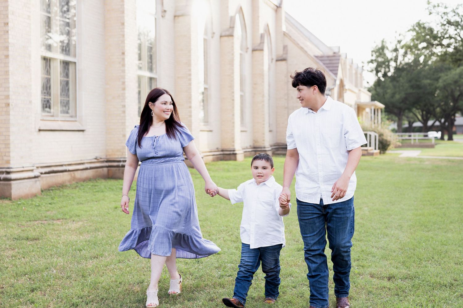 Pregnant woman in blue dress walks holding hands with husband and young son on green lawn.