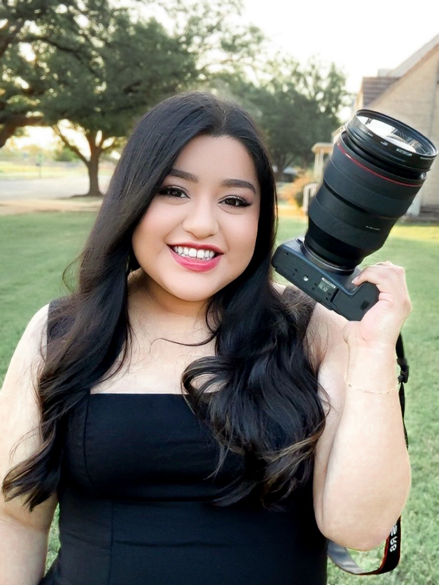 A smiling young woman holds a professional camera on her shoulder outdoors.