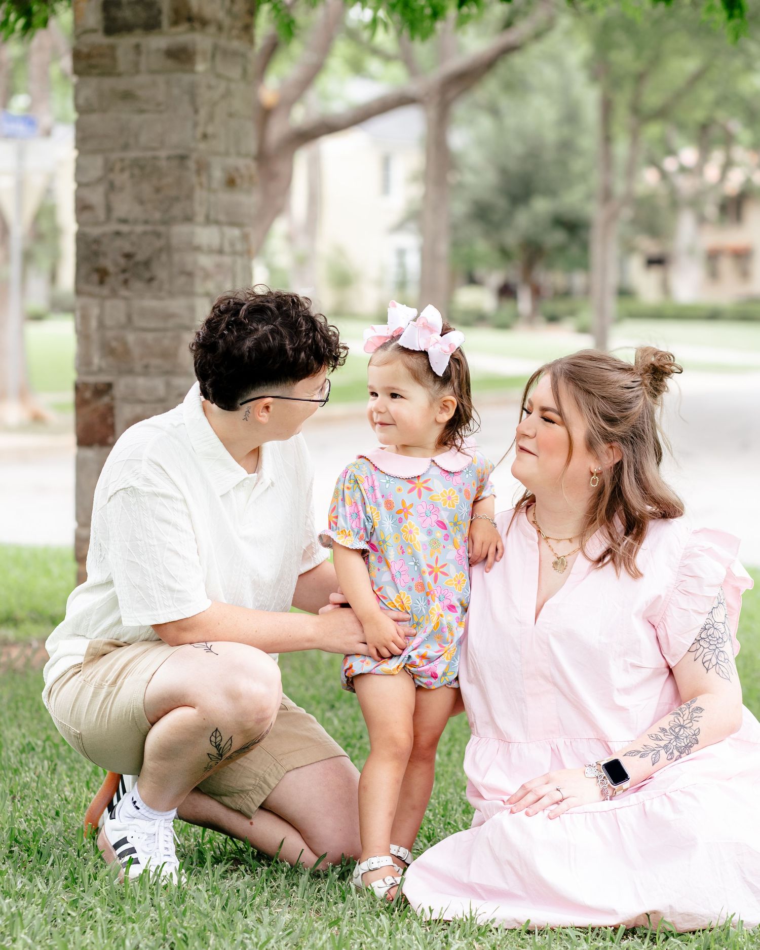 Family of three poses outdoors on grass, parents smiling at toddler girl in colorful dress with pink bow.