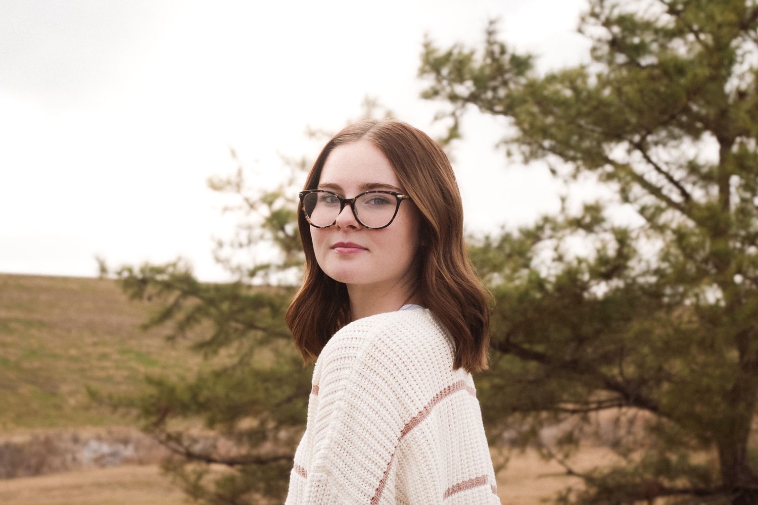 Young woman with glasses and brown hair wearing a cozy sweater, posing outdoors near green trees.