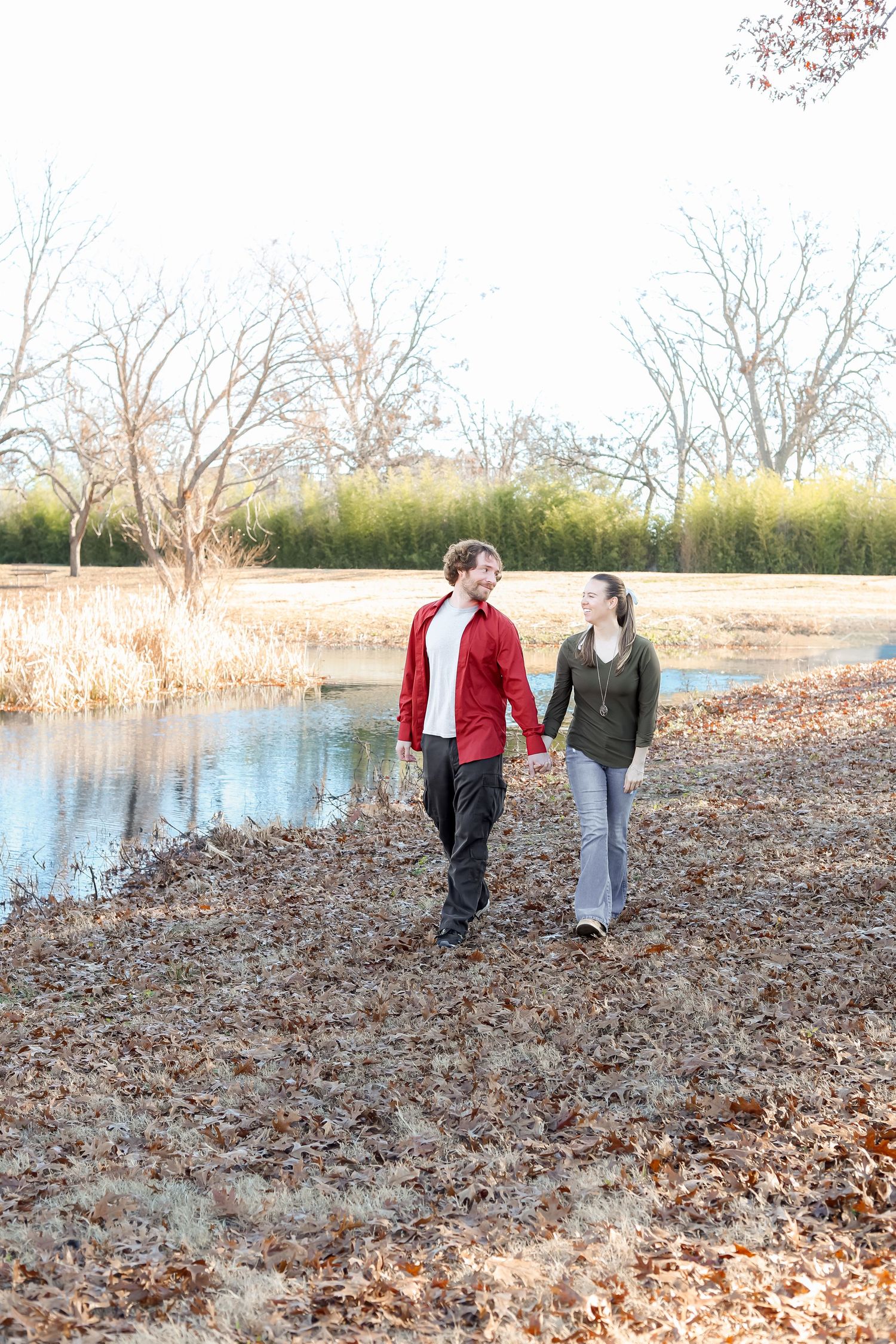 Young couple holding hands walking along a pond surrounded by autumn leaves and bare trees.