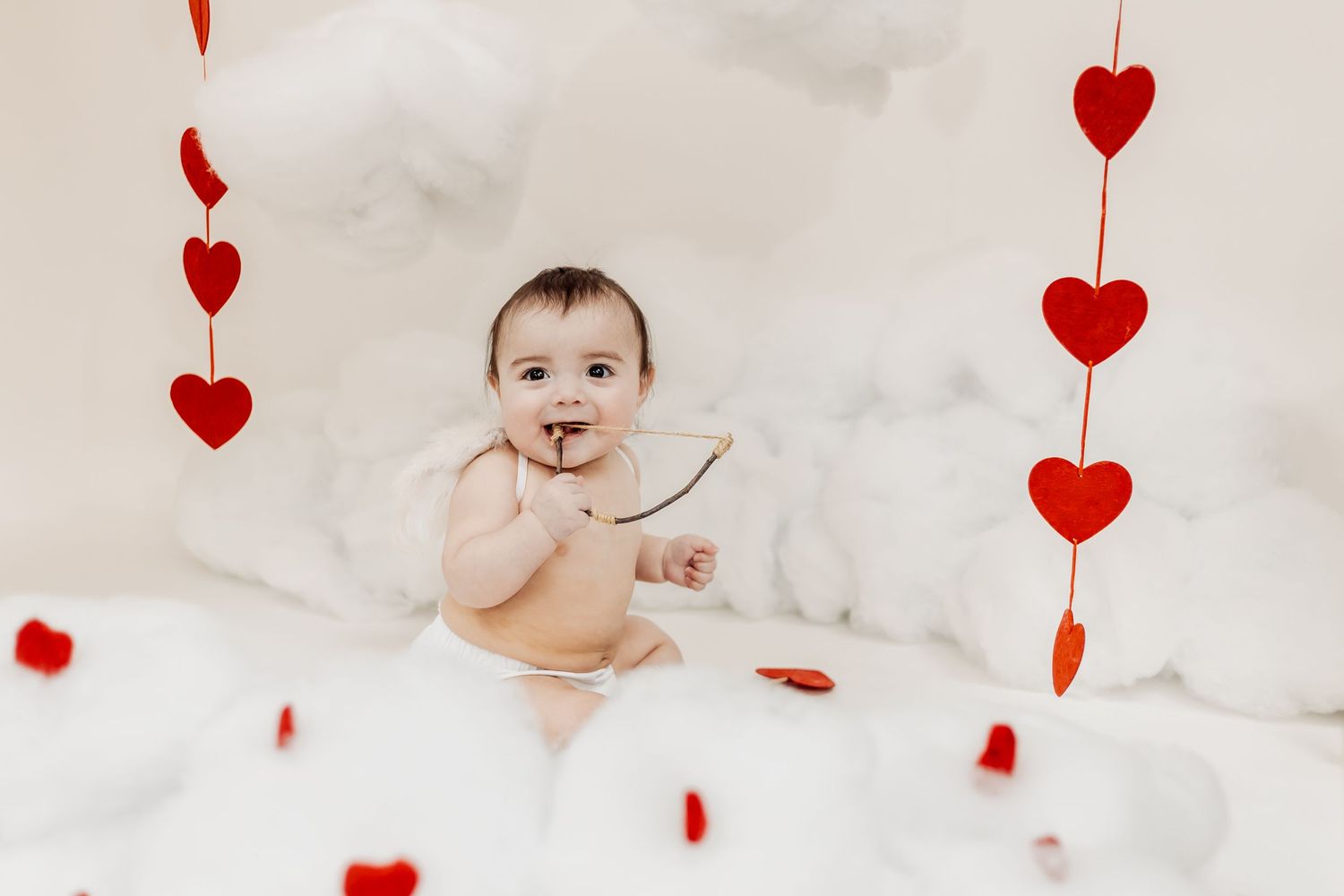 Baby sitting among white fluffy clouds with red heart garlands for a Valentine's Day themed photo shoot.