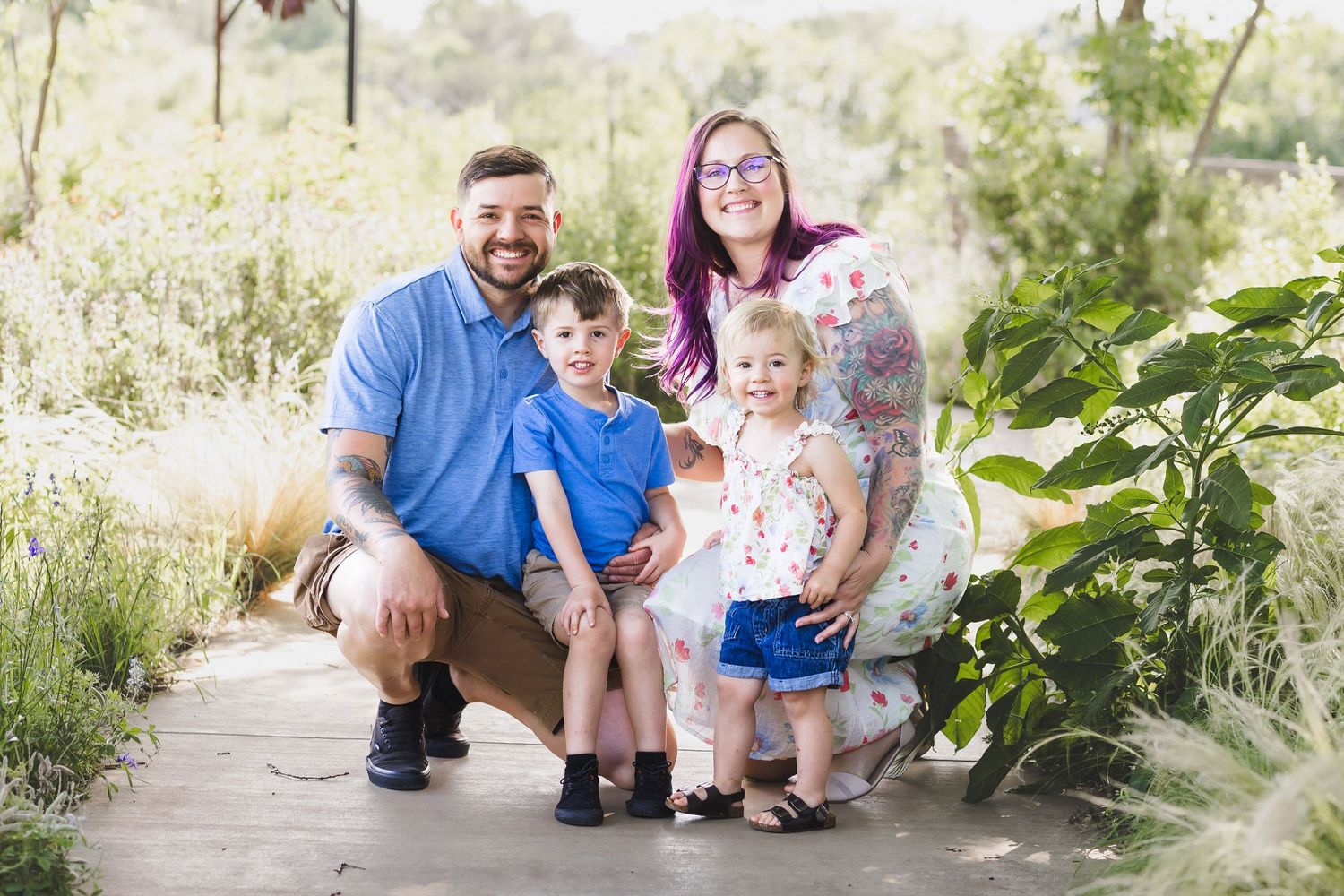 Family of four smiling outdoors on a wooden path surrounded by lush greenery, parents with two young children posing together.