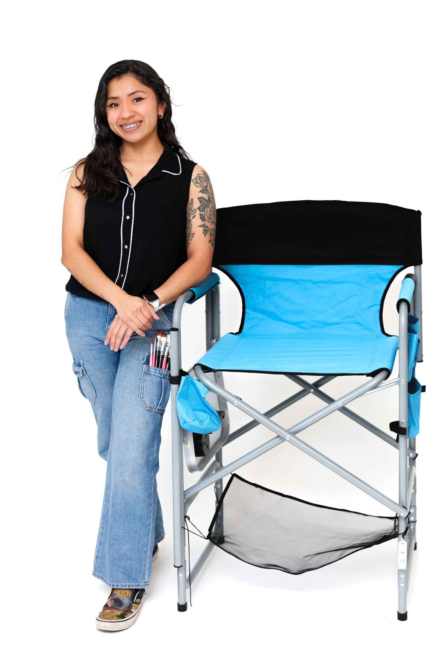 A smiling tattooed woman leans on a blue folding director's chair against a white background.