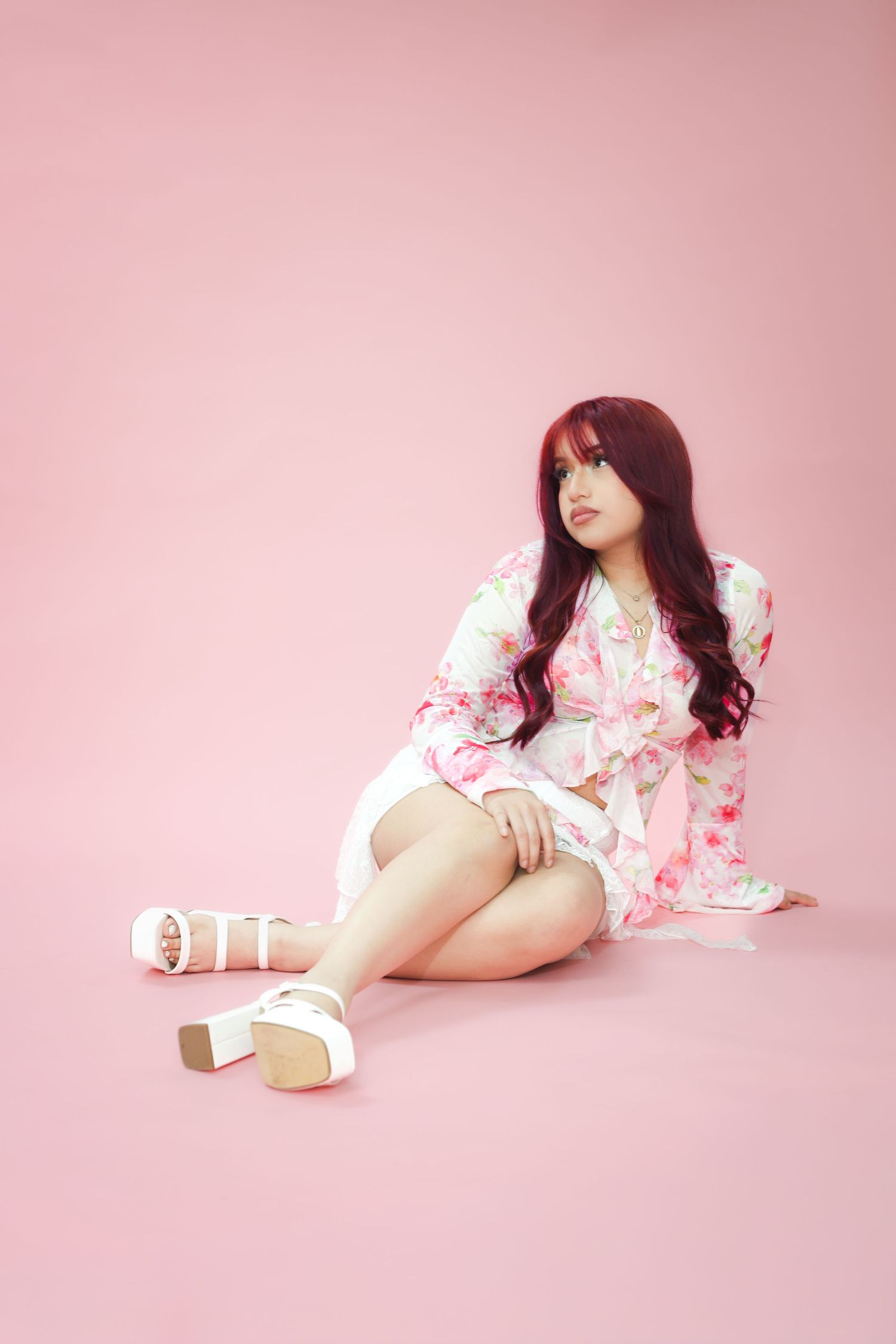 Young woman with red hair sitting on pink background wearing floral kimono and white platform sandals.