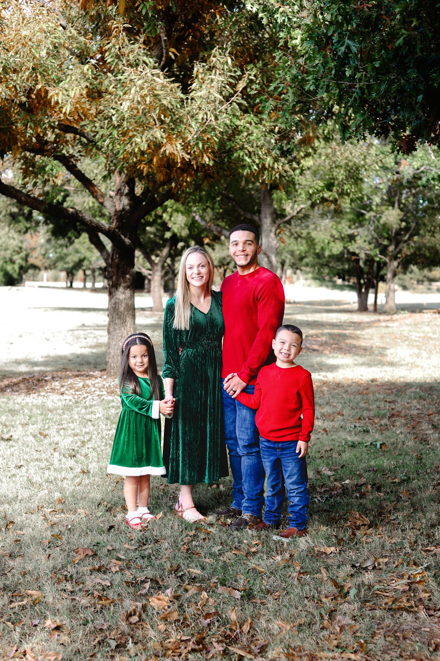 Happy family of four in coordinated red and green Christmas outfits posing under trees in a park.