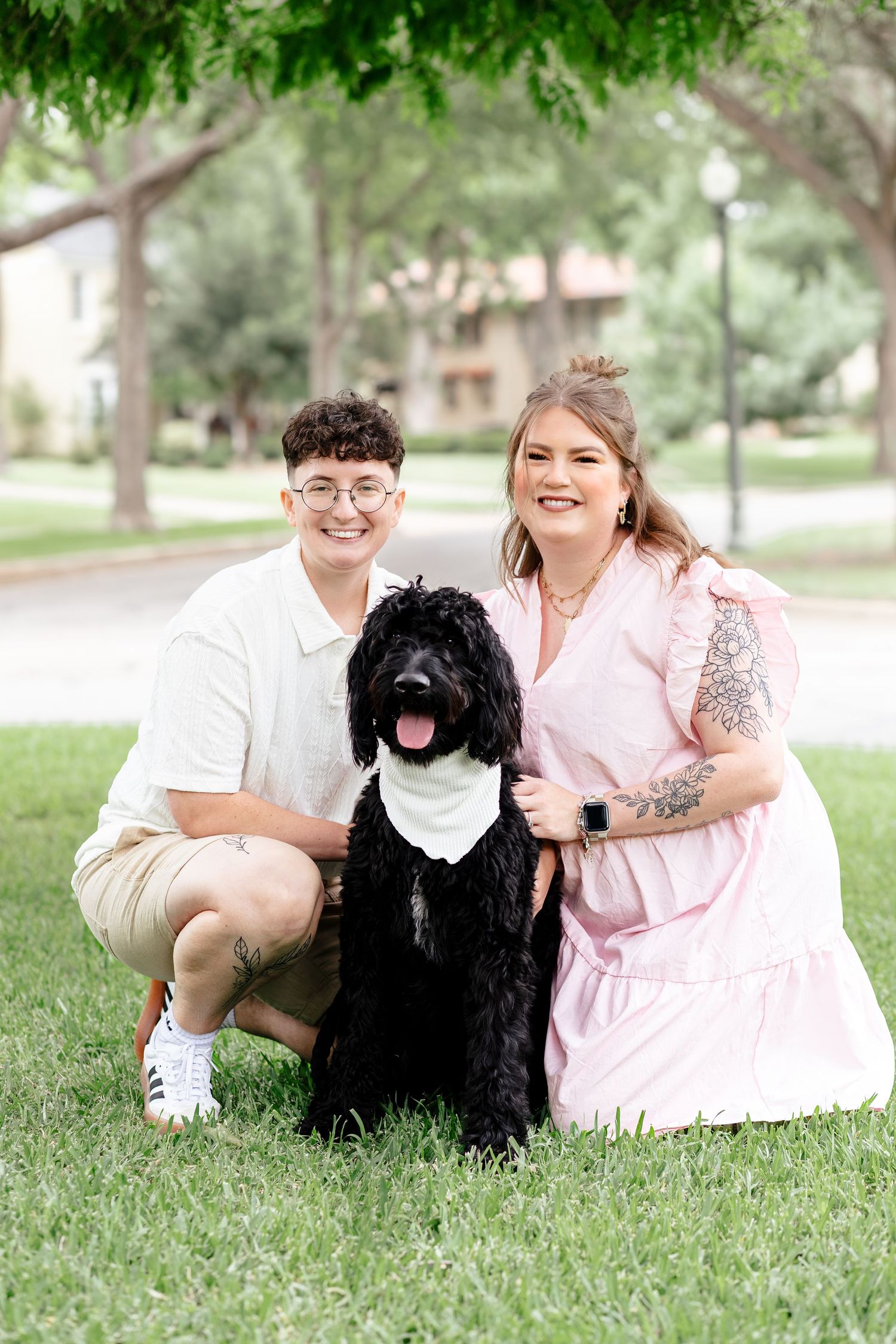 Couple smiling with black doodle dog wearing bandana on green grass in sunny park setting.