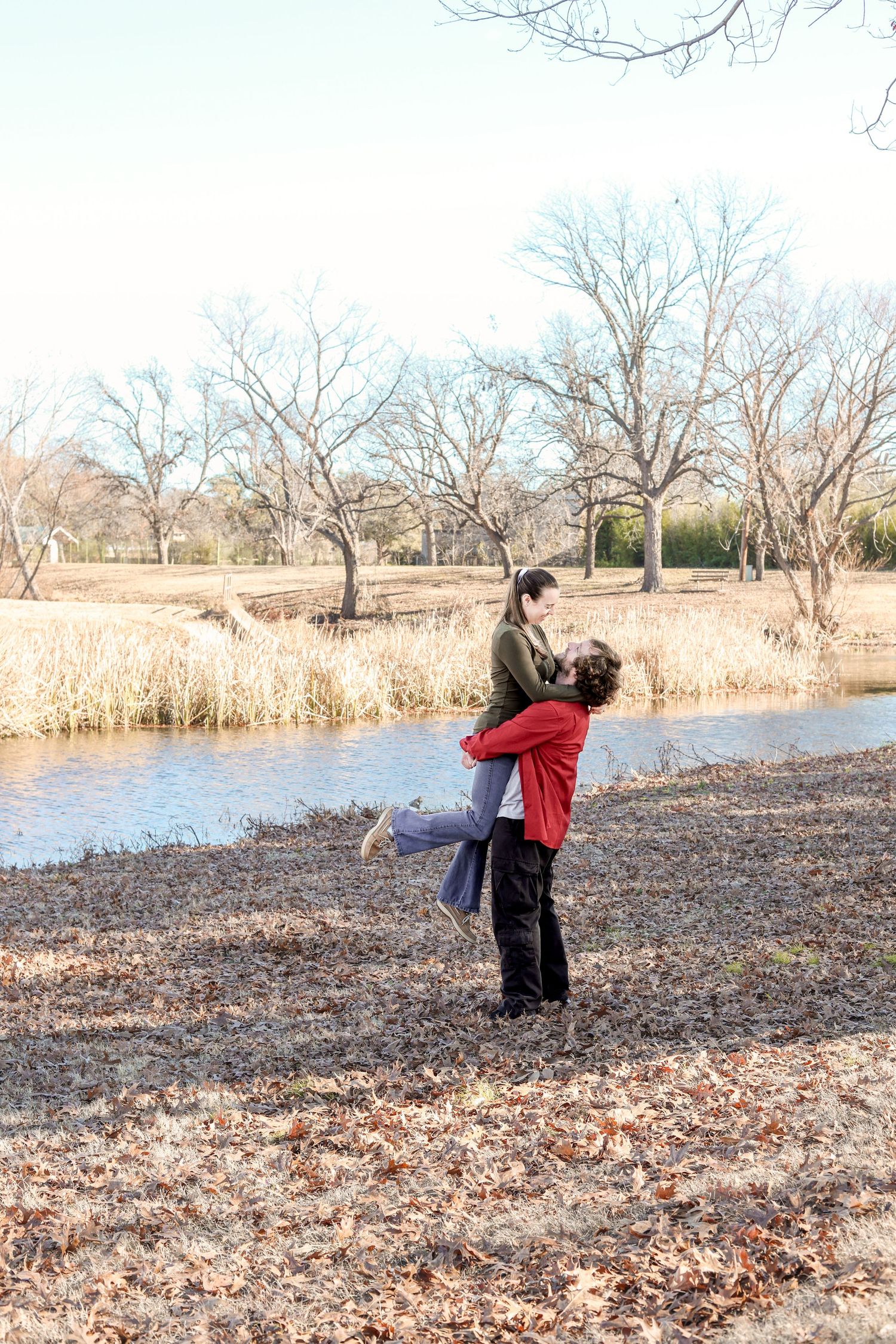 Couple sharing a romantic moment by a serene pond in a bare winter park, man lifting woman joyfully.