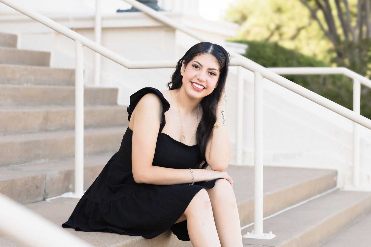 Young woman in black dress smiling on outdoor steps, long dark hair, casual portrait photography.