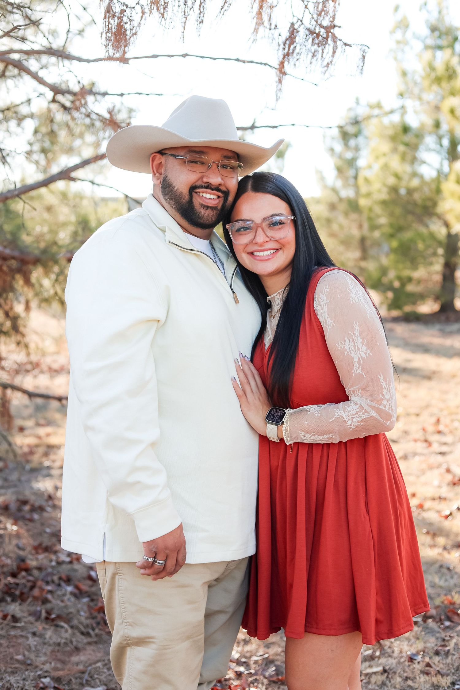 Happy couple embracing outdoors, man wearing a white blazer and cowboy hat, woman in a red dress.