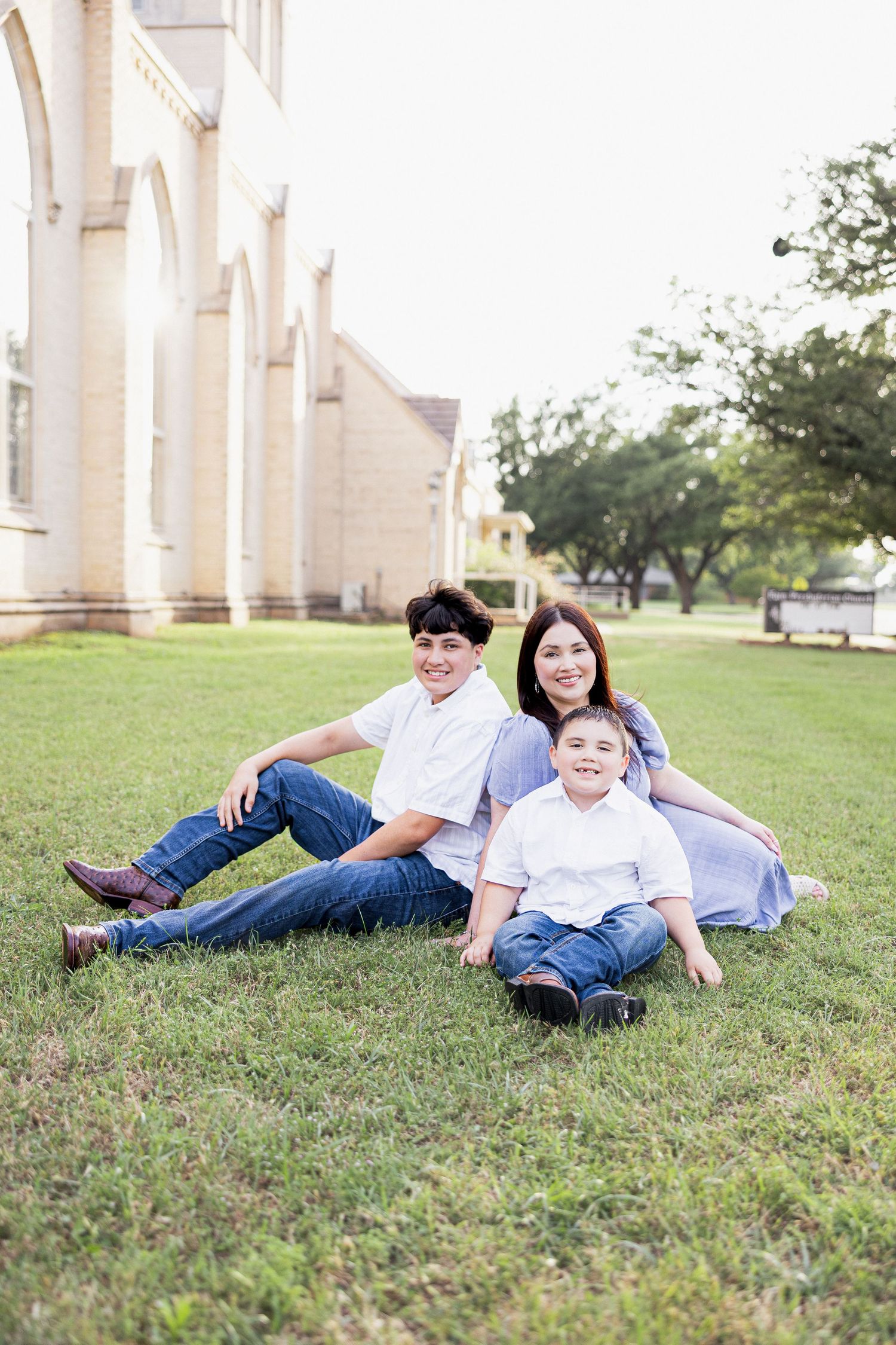 Mother in blue dress sits on grass with two smiling sons near a historic building outdoors.