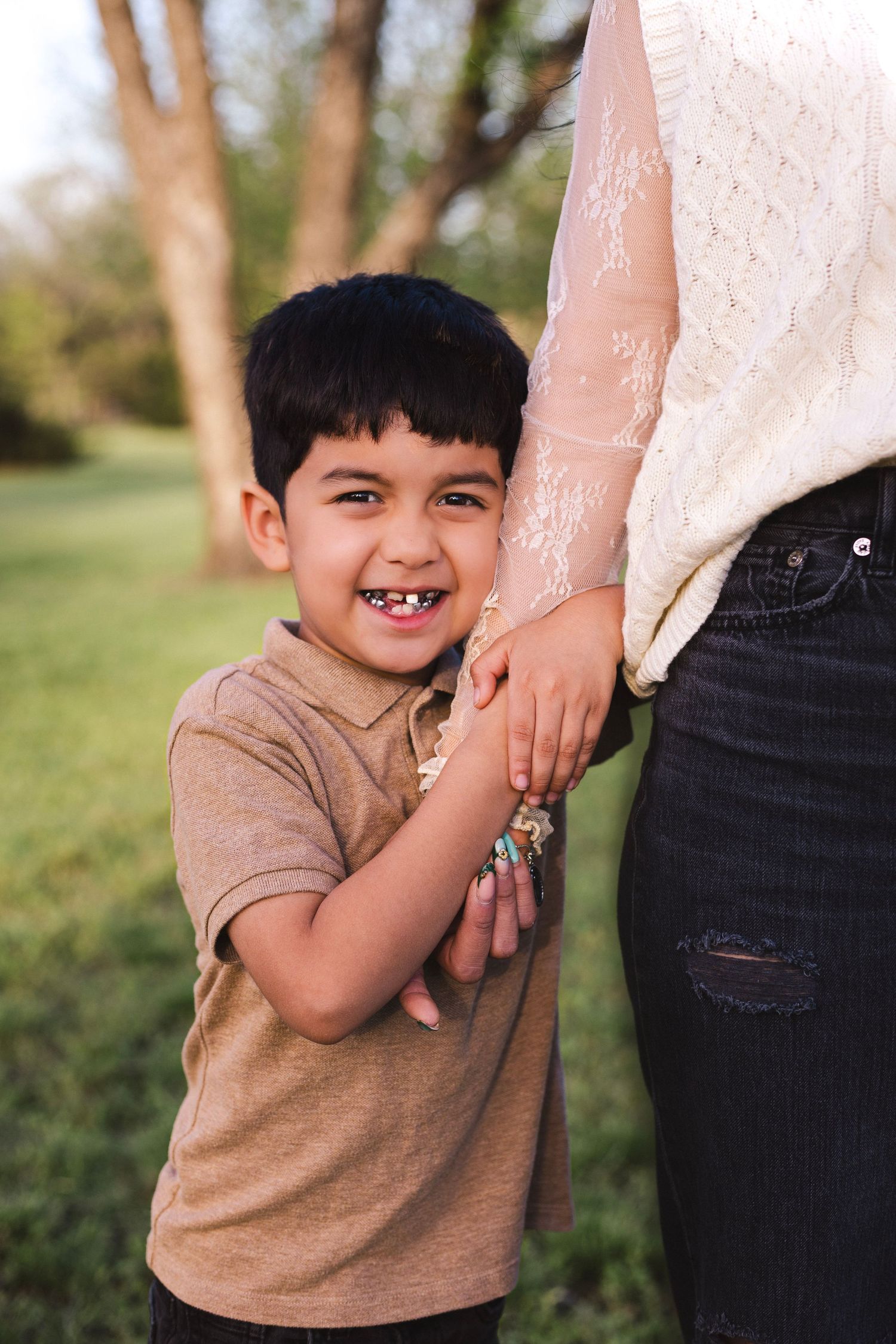 A happy young boy hugs his mothers hand while smiling brightly in an outdoor park setting.