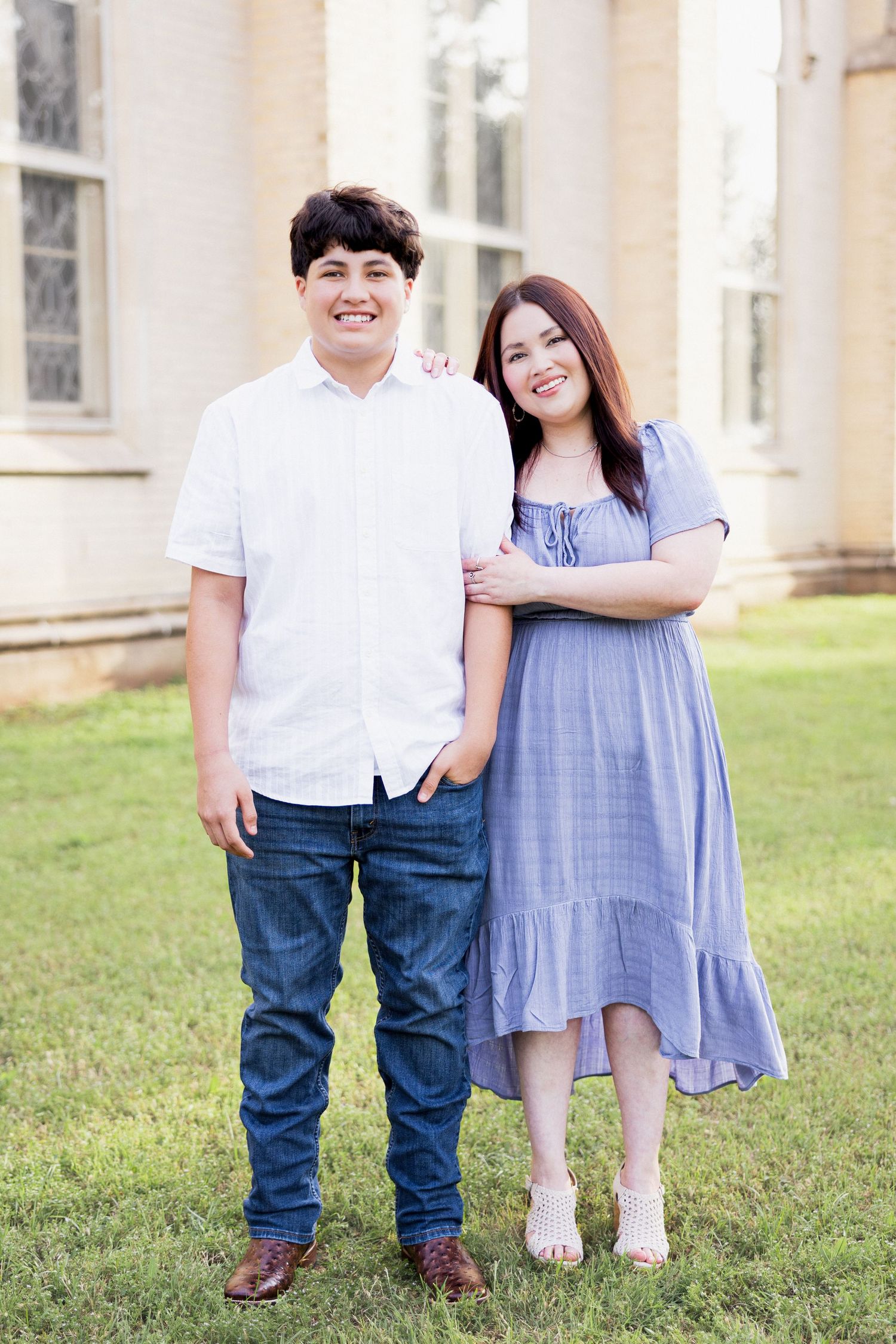 Smiling teenage boy and girl in blue dress pose together on green lawn near a classic building.