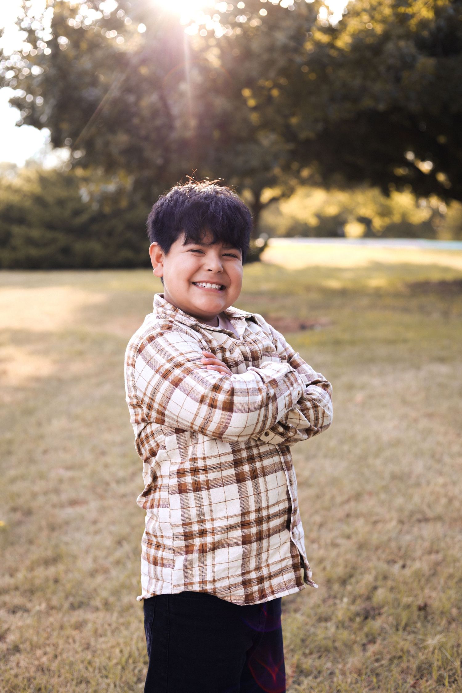 Smiling boy with arms crossed wearing plaid shirt in sunny outdoor park setting.